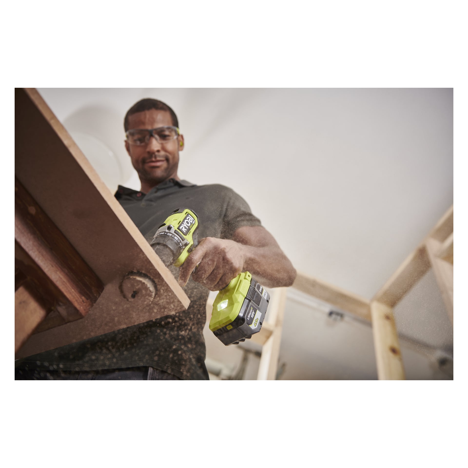 Eine Person mit Sicherheitsbrille bohrt drinnen mit einem Ryobi One+ HP Brushless-Bohrer ins Holz, während Sägemehl fliegt. Zwei Batterien sichtbar.