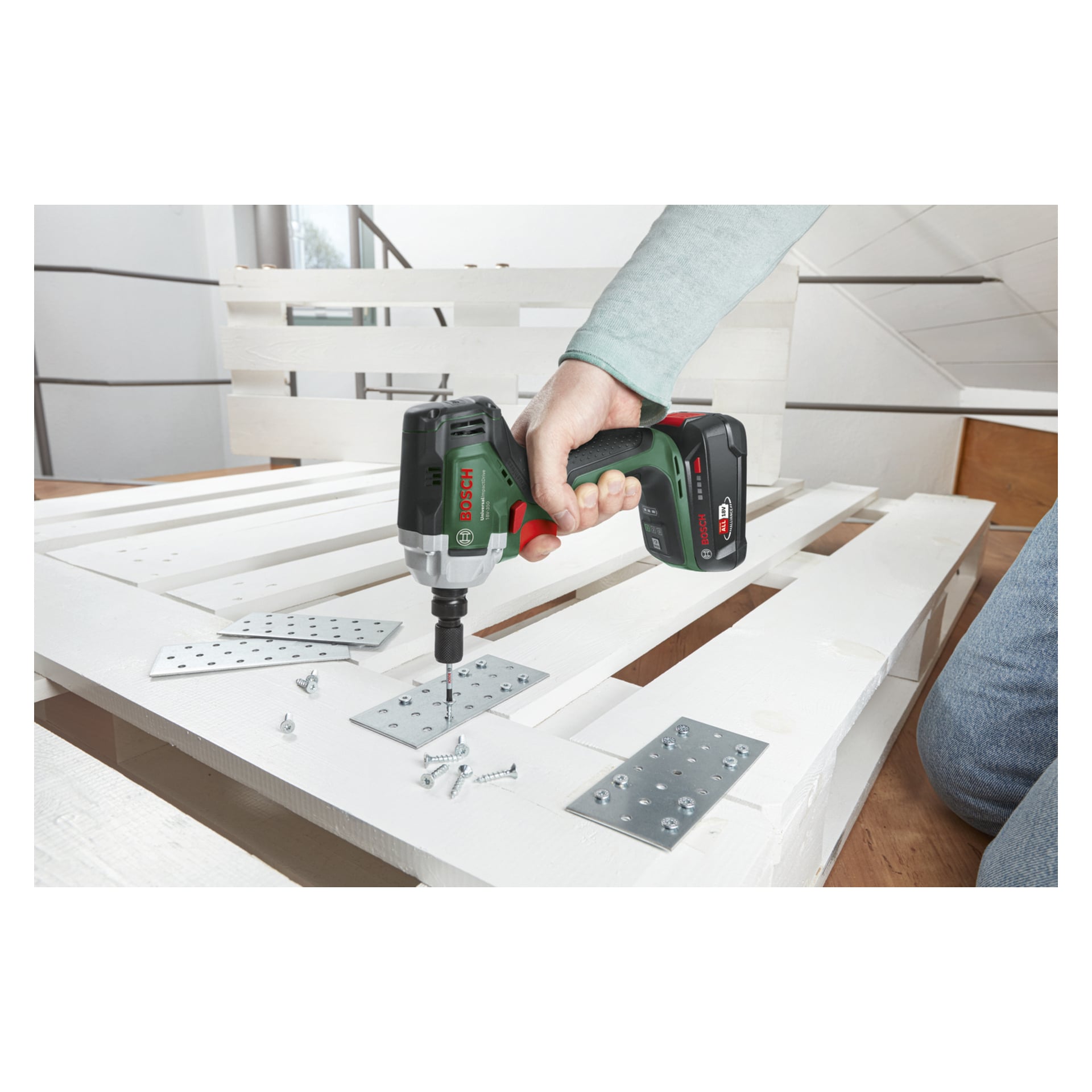 Osoba mocuje metalową płytkę do białej drewnianej palety za pomocą Bosch UniversalImpactDrive 18V-350; W pobliżu leżą śruby i płyty.