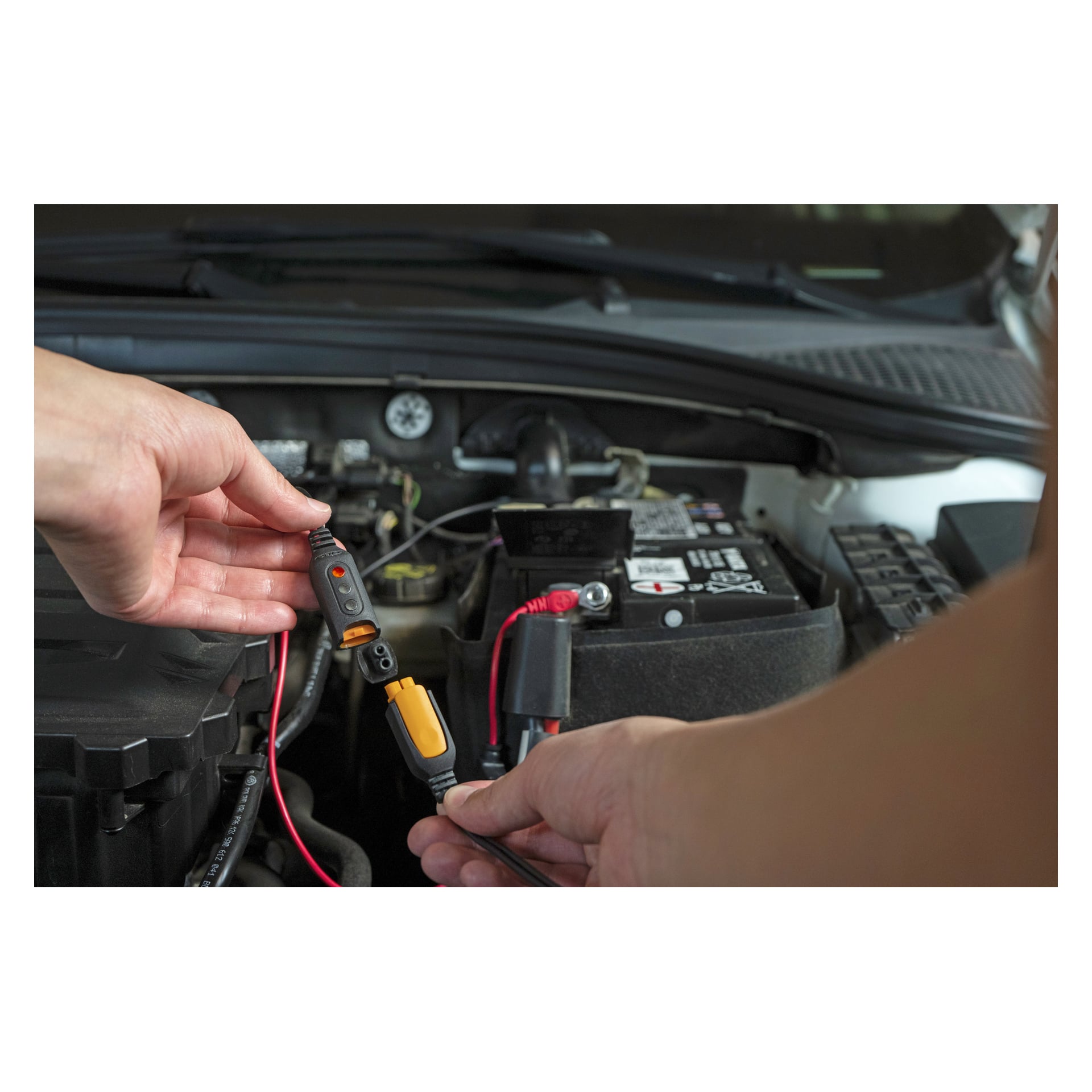 Händerna fäster en CTEK MXS 5.0 Power Kit-laddare och powerbank på bilbatteripoler under den öppna huven, motordelar bakom.
