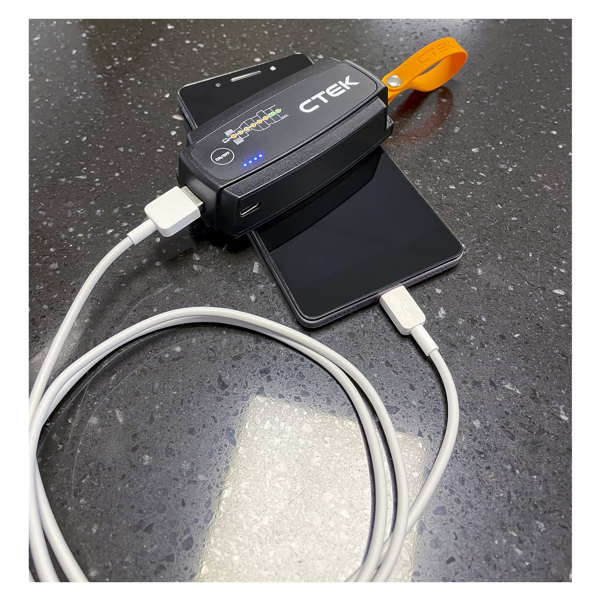 Ett CTEK MXS 5.0 Power Kit med indikatorlampor och orange rem laddar en smartphone; En annan telefon ligger nedanför på en mörk bänk.