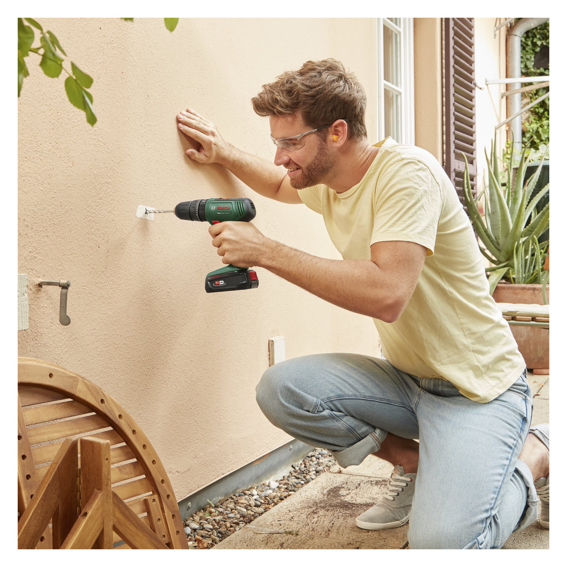 En mann med vernebriller og ørepropper kneler utendørs og borer et hull i en beige vegg med en Bosch batteridrevet bormaskin.