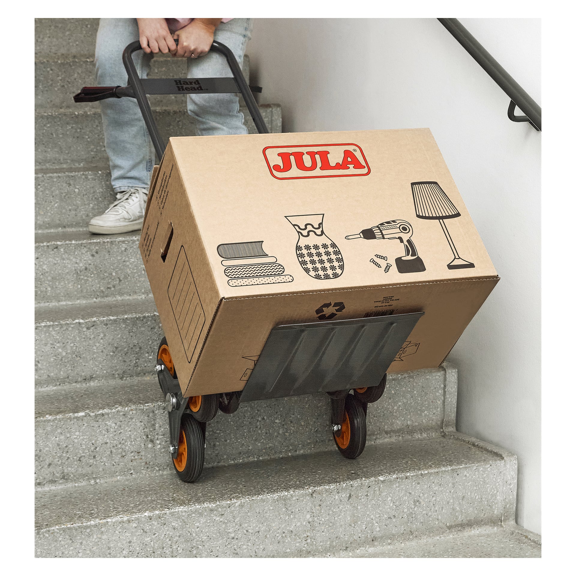 Osoba przenosi duże pudełko JULA pokryte ikonami przedmiotów domowych po dywanach po schodach za pomocą Hard Head Stair Climber Barrow.