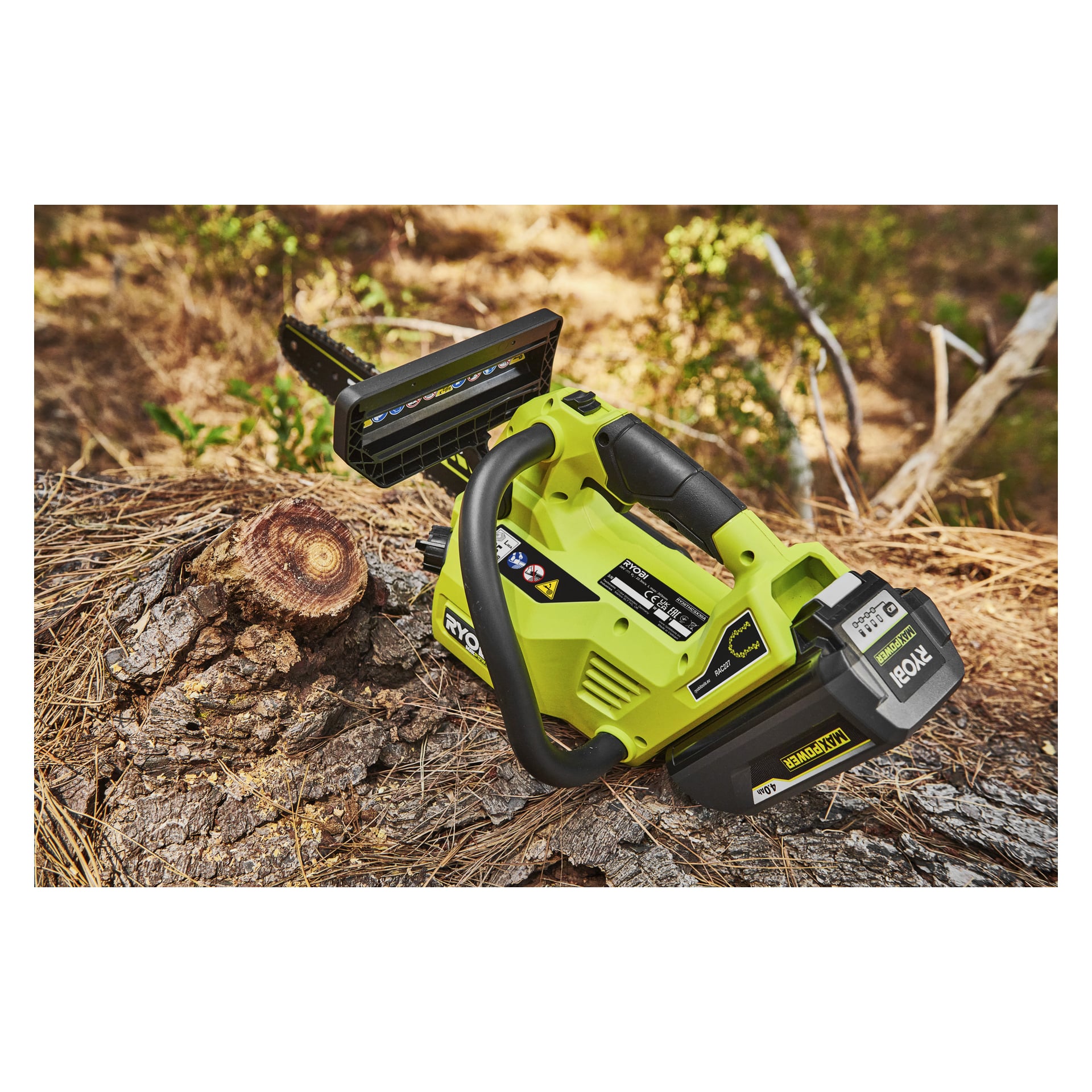 En Ryobi Max Power-batterimotorsåg RY36THCSX30A-0 vilar på en stubbe bland tallbarr, med torrt gräs och grönska bakom.
