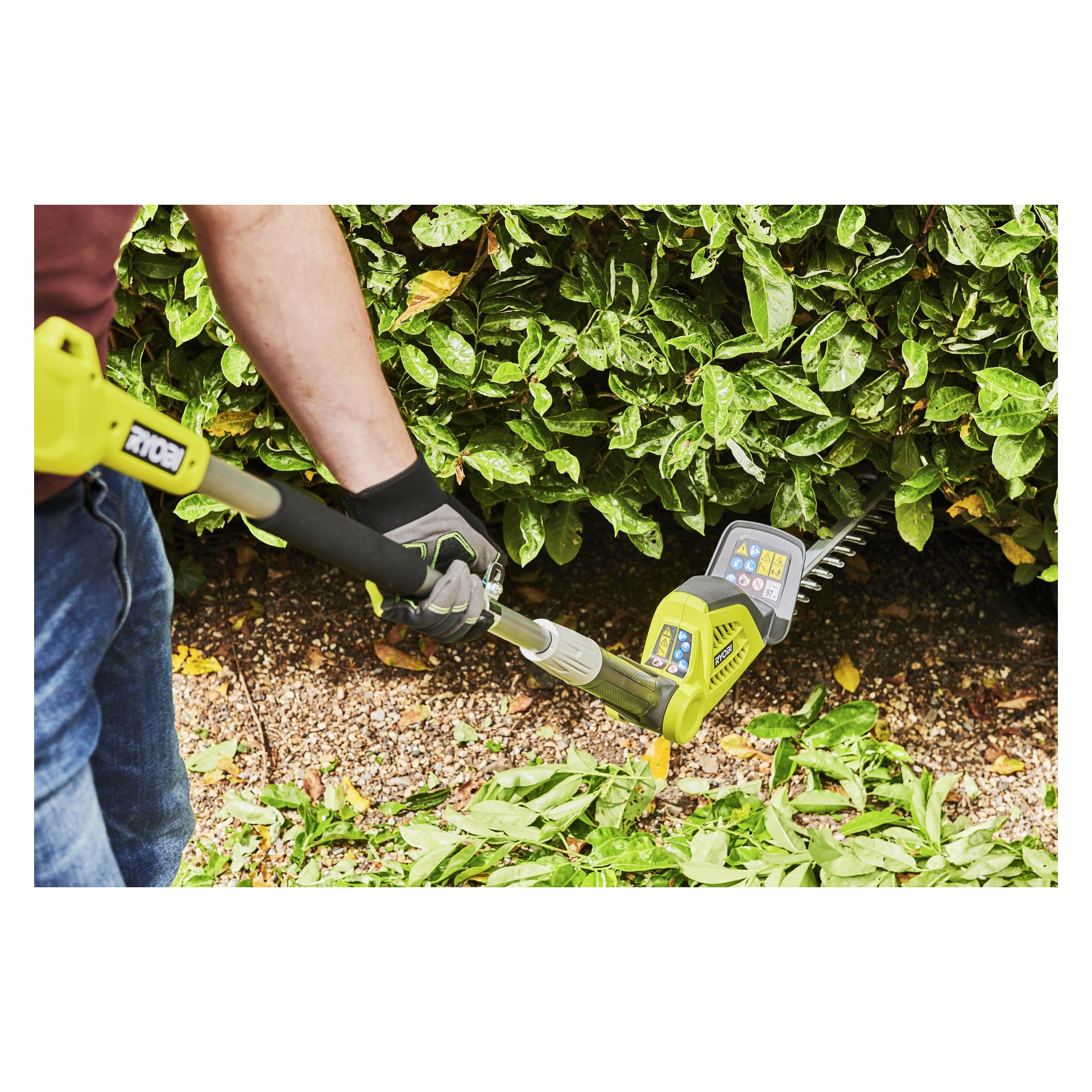 En person med hansker bruker en Ryobi Max Power Battery hekksaks RY36PHT50A-0 for å forme busker, med klipp på bakken.