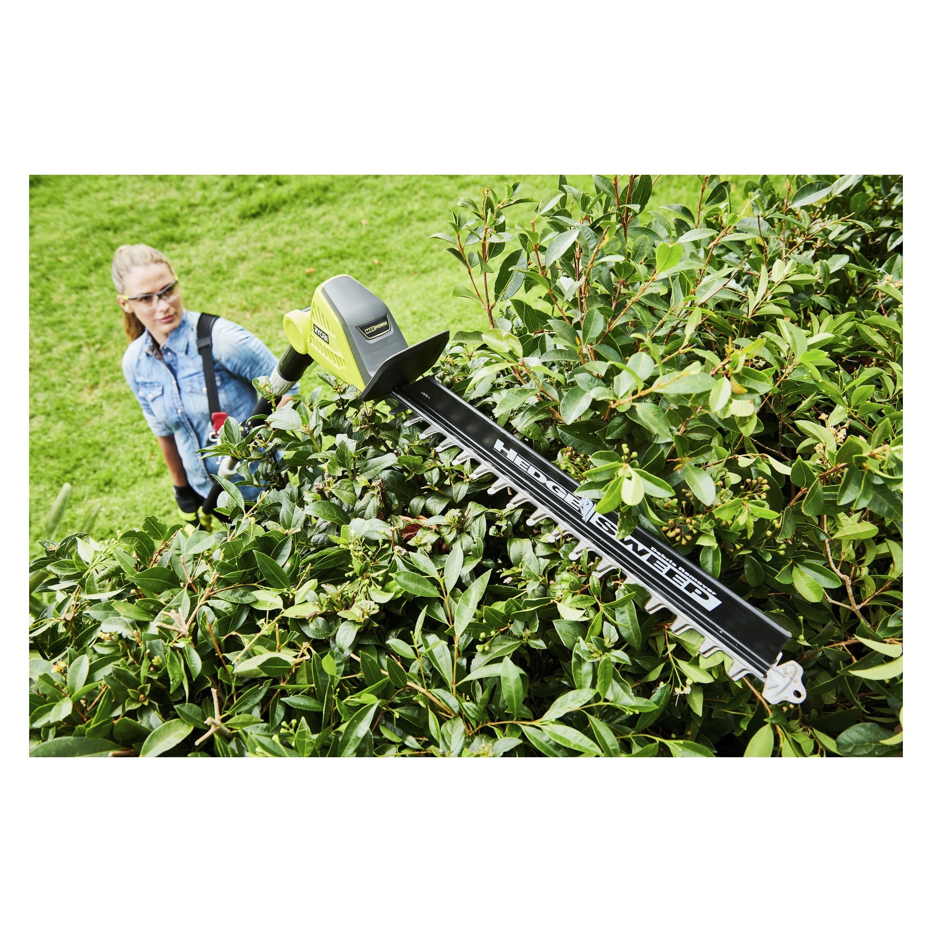 En person med vernebriller og hansker trimmer tette grønne busker utendørs med en Ryobi Max Power trådløs hekkesaks, holdt over hodet.