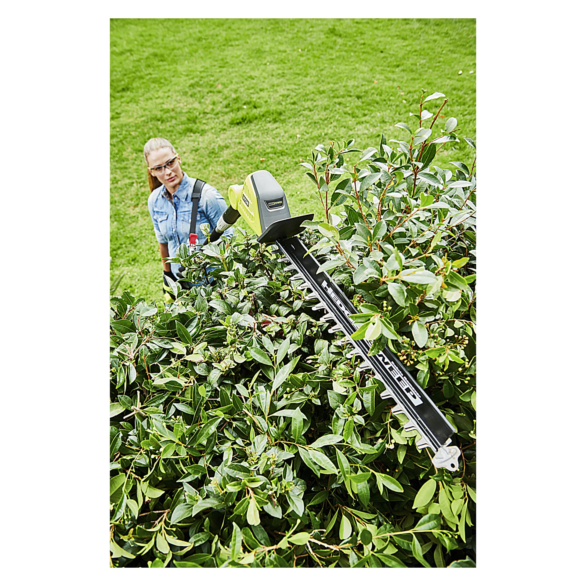 En person med vernebriller trimmer en høy grønn hekk utendørs med en Ryobi Max Power Battery hekkeklipper RY36PHT50A-0.