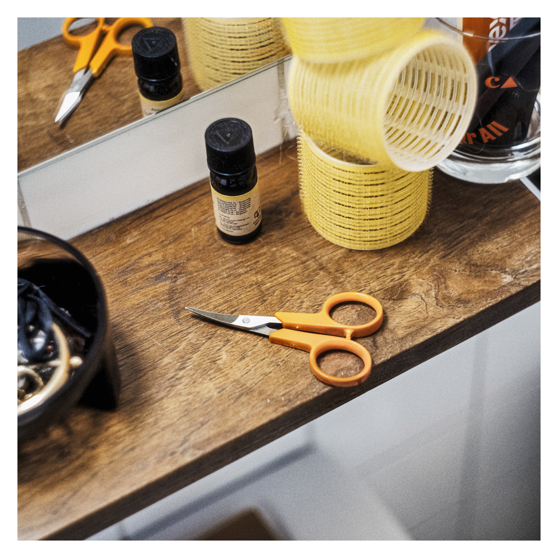 Fiskars neglesakser med oransje håndtak, gule hårruller, en svart beholder, en liten flaske og glasskopp på tre ved et speil.