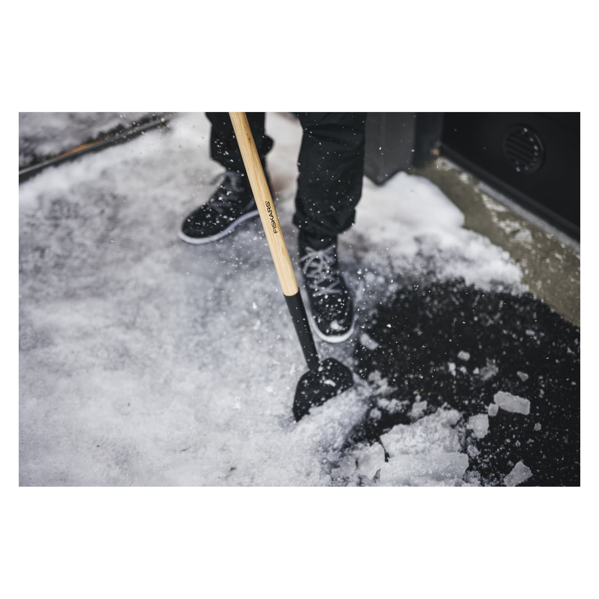 En person i svarta byxor och skor använder en Fiskars Ice/barking iron Classic med ett trähandtag för att bryta isen och sprida isbitar.