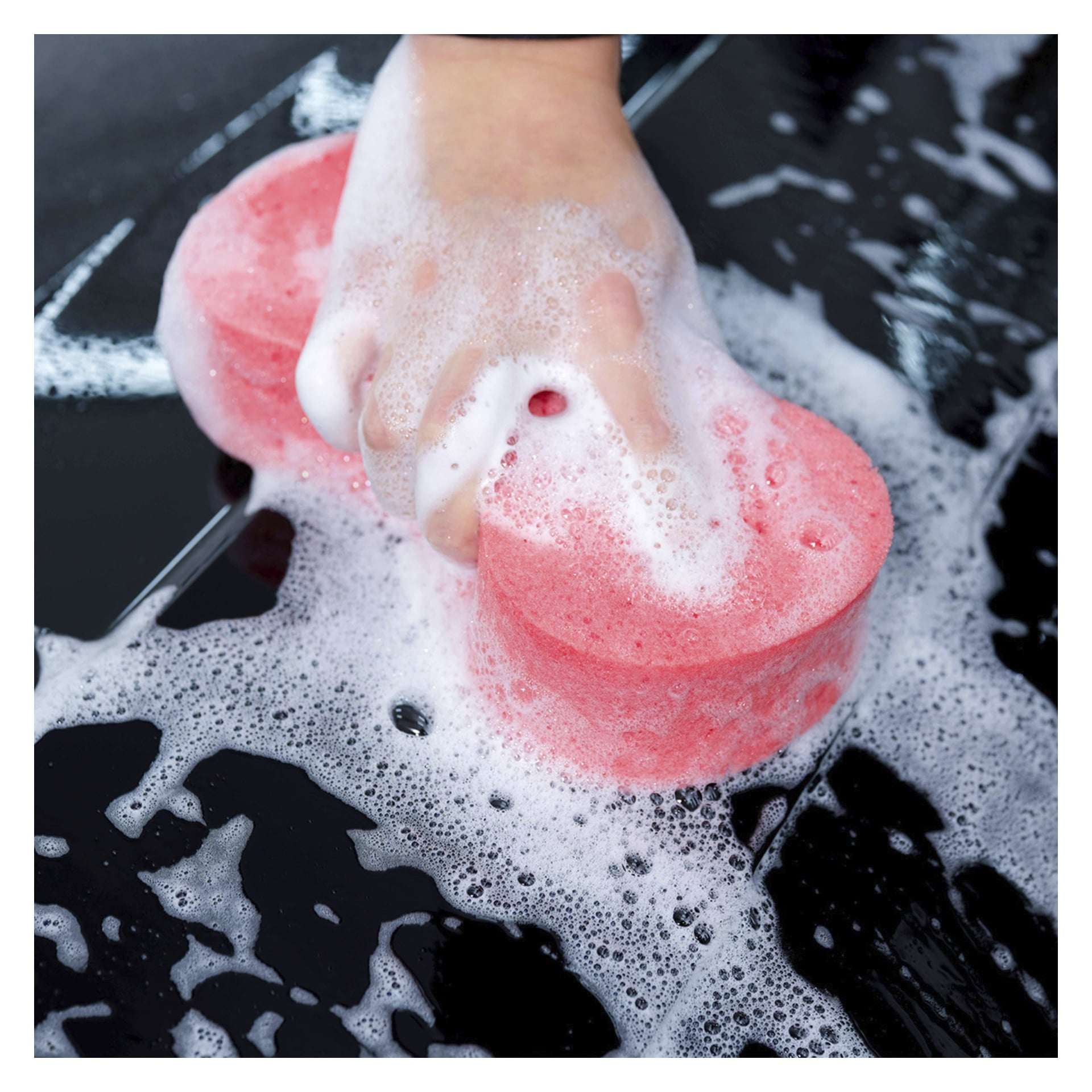 En hand rengör en svart yta med en rosa, tvålig svamp från "Sonax Car care kit 8 pieces." Bubblor och vatten finns runt svampen.