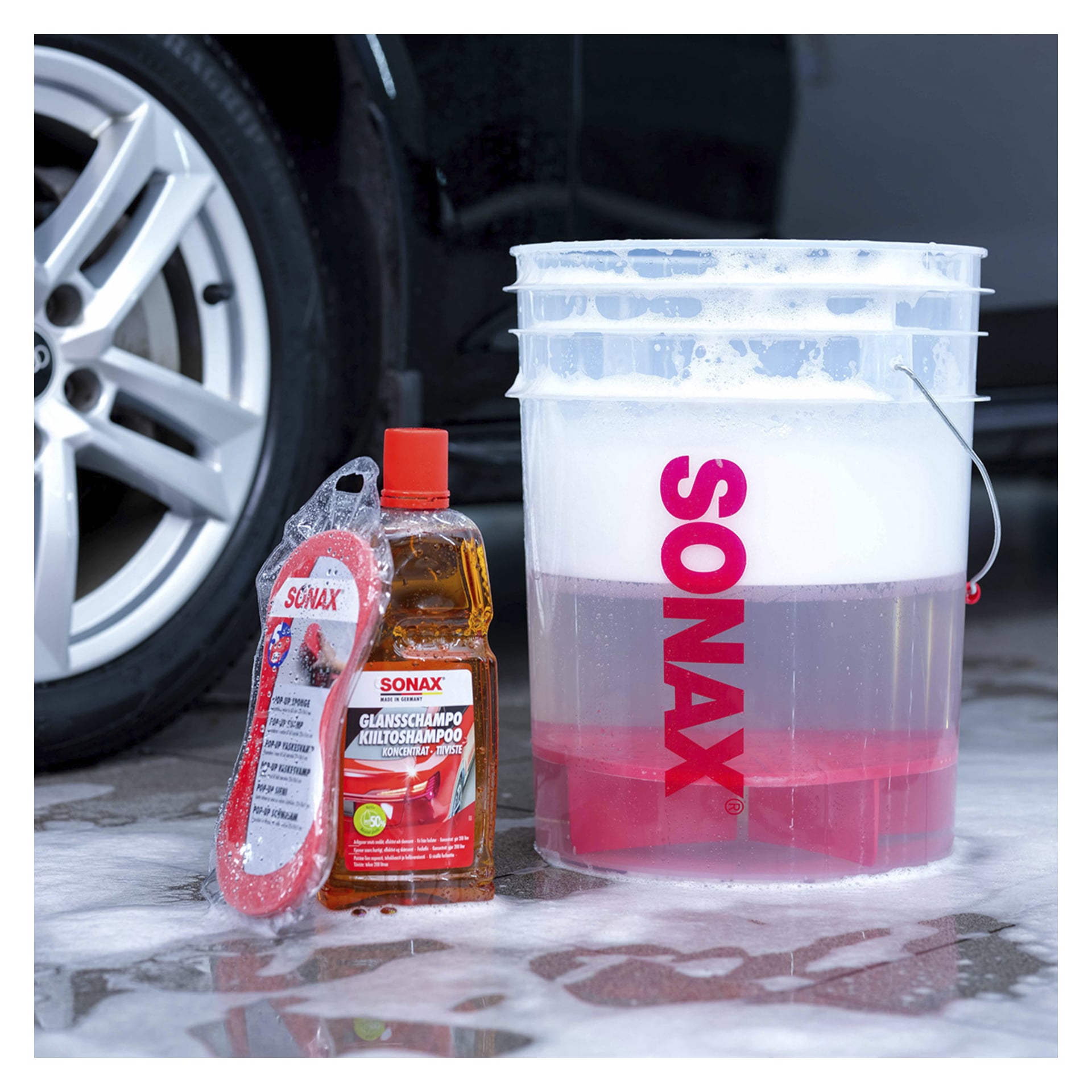 En klar hink märkt "Sonax" fylld med tvålvatten står på våt asfalt, bredvid en "Sonax Car care kit 8 pieces" och en röd rengöringssvamp. Ett bilhjul syns i bakgrunden.