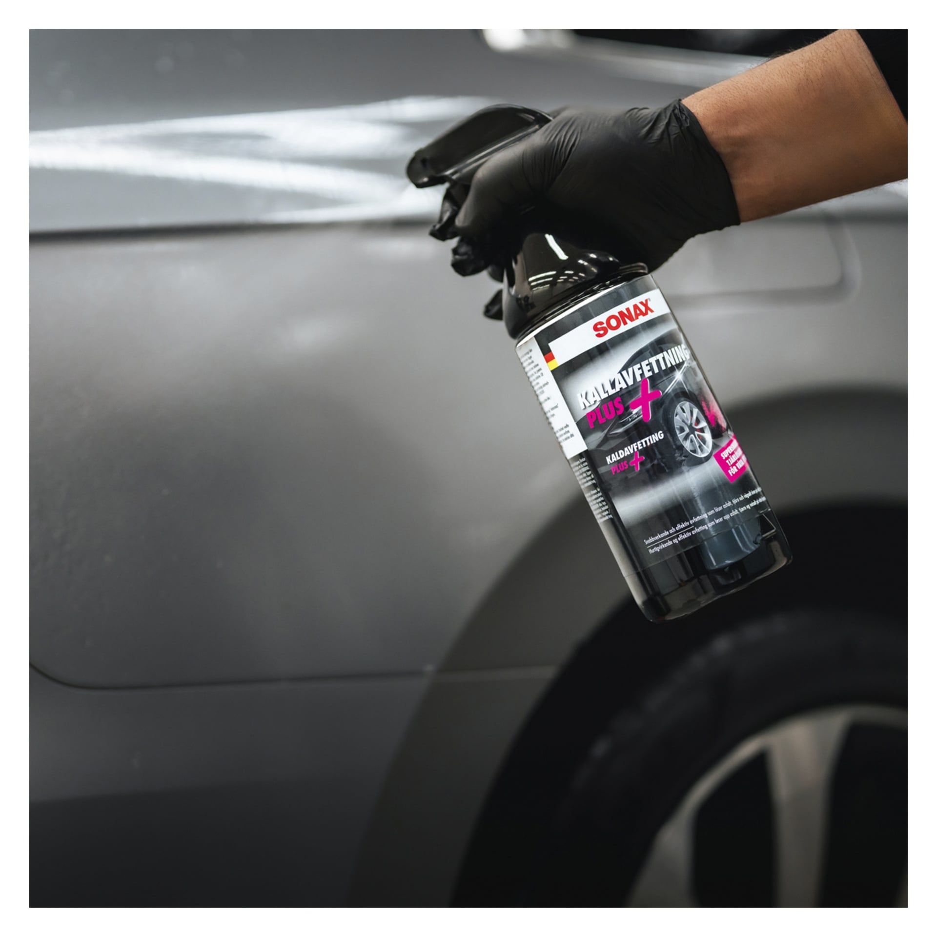 En handske sprayar ett Sonax Car Care Kit 8-delat produkt på sidan av en mörk bil, med fokus på området nära bakhjulet och dörren.