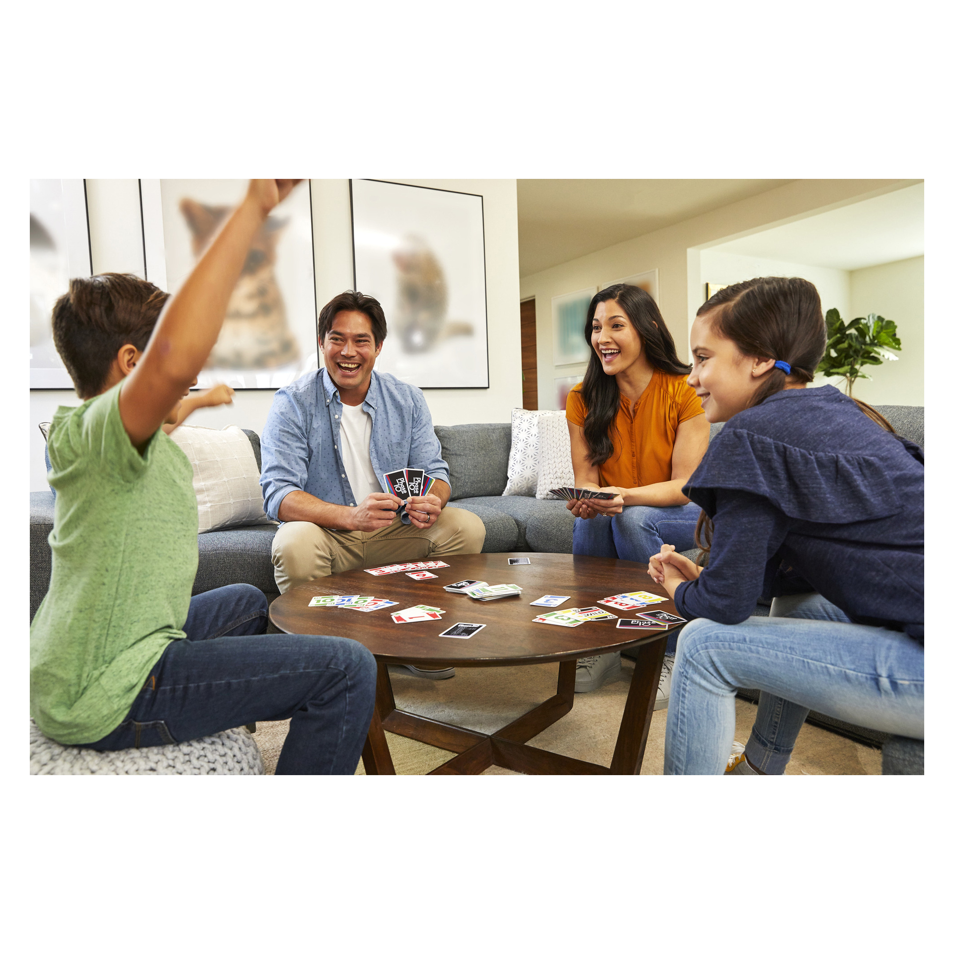 Fyra familjemedlemmar spelar UNO vid ett soffbord, leende i ett ljust och mysigt vardagsrum med väggkonst och utspridda kort.