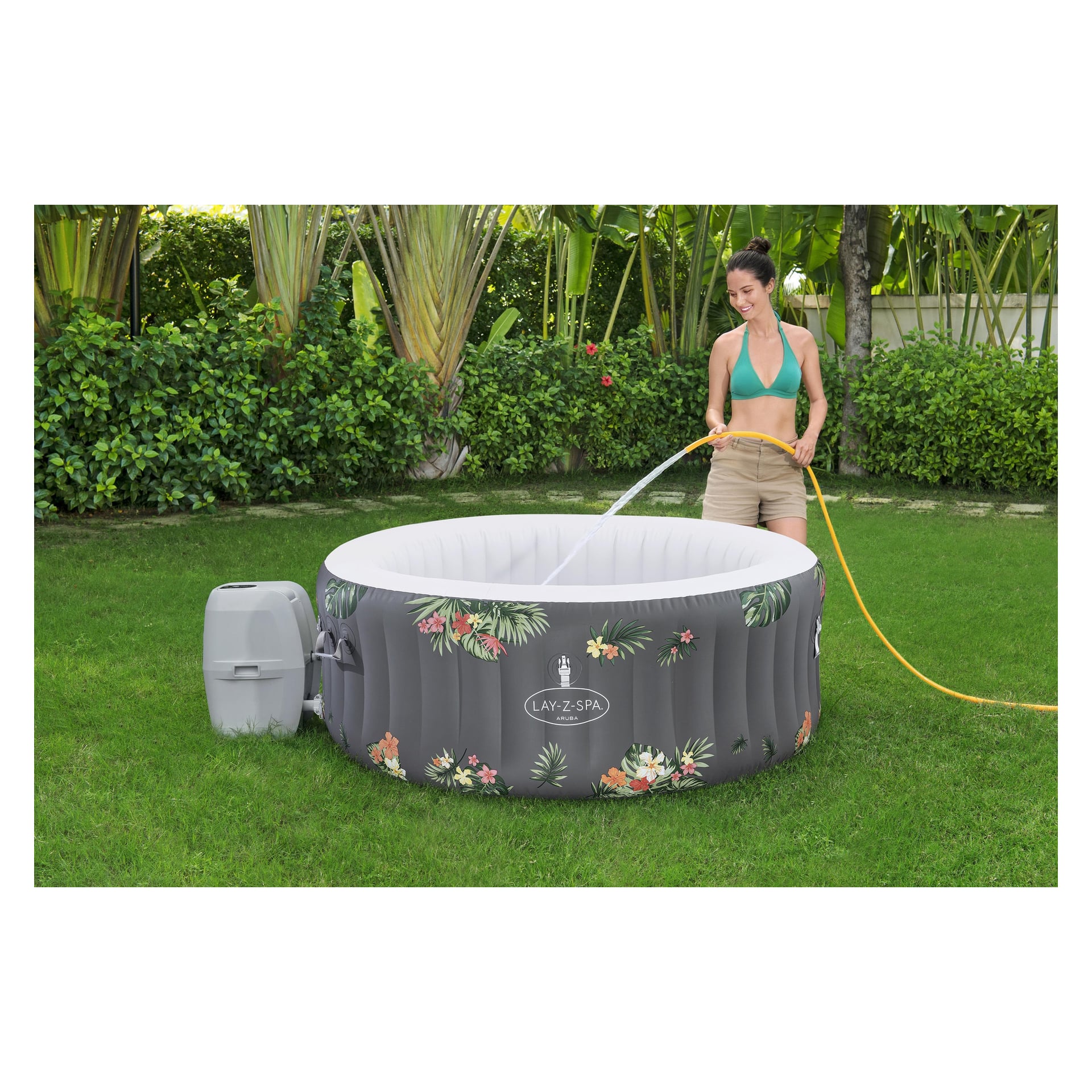 Kobieta w zielonym topie bikini i beżowych szortach używa żółtego węża, by napełnić Bestway LAY-Z-SPA Aruba AirJet na trawiastym trawniku z palmami.