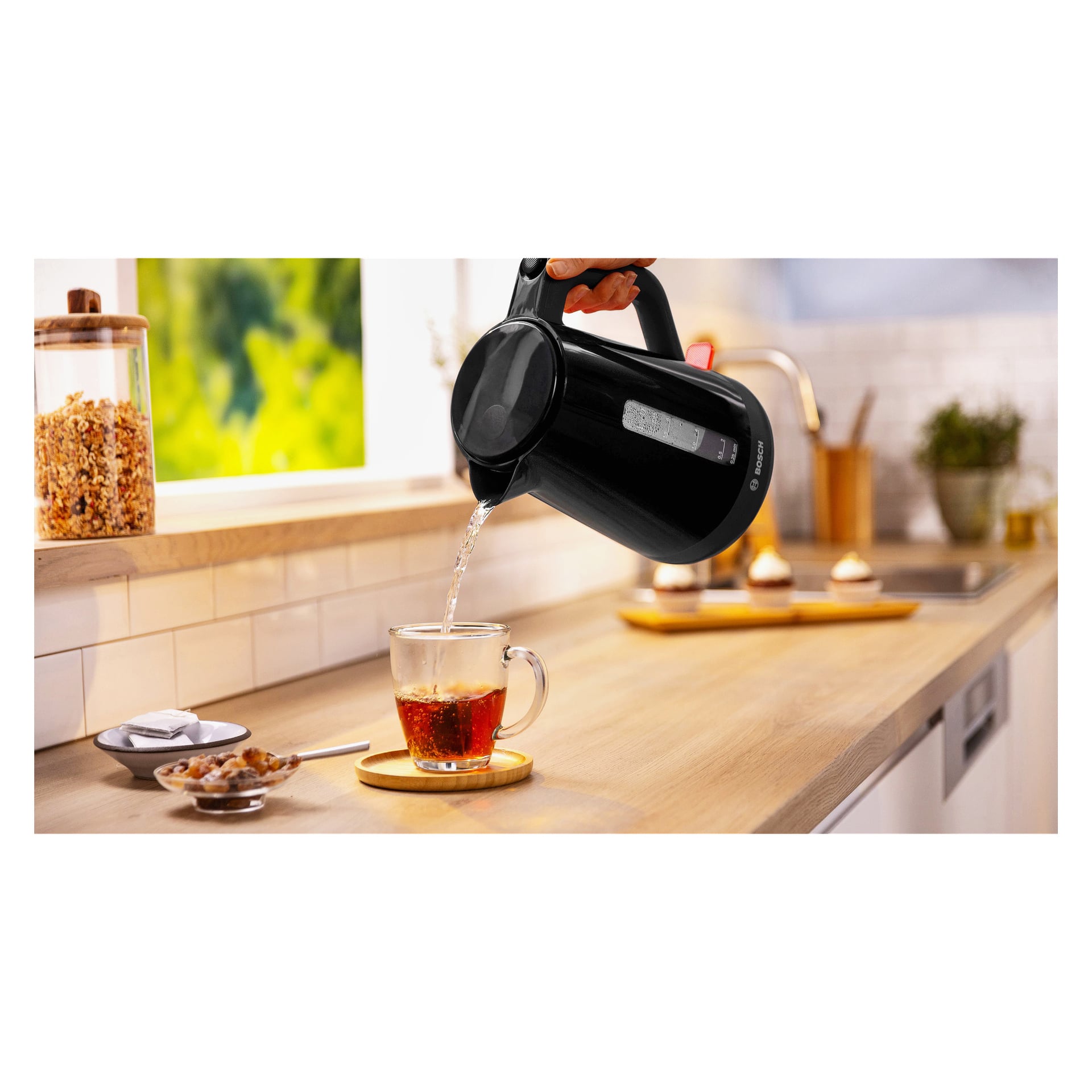 Varmt vann helles fra en Bosch Home Kettle i en glasskopp med te på kjøkkenbenken med glass, boller og cupcakes bak.