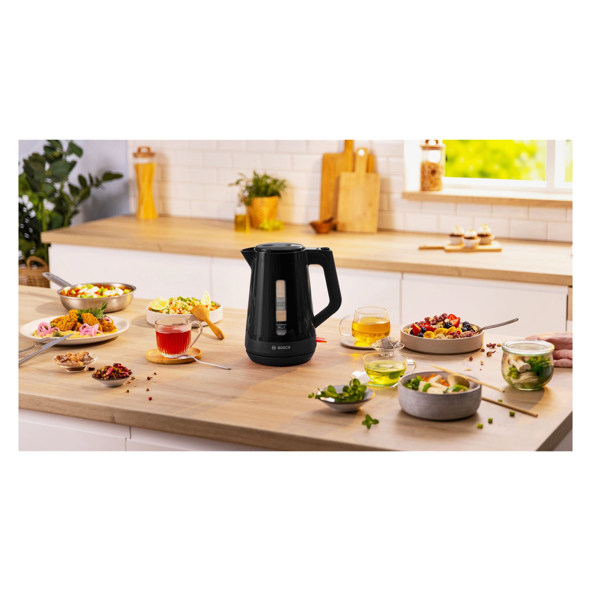En svart Bosch Home Kettle MyMoment TWK1M123 står på en trebenk med mat, tekopper og tallerkener; Planter på vinduskarmen.