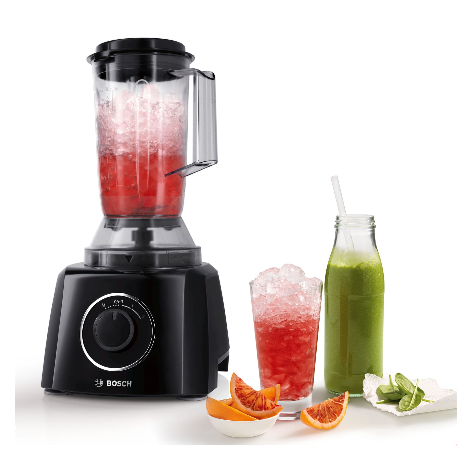 En svart Bosch matprosessor med rød væske og is står ved siden av et glass, en grønn juiceflaske og boller med appelsin- og limeskiver.