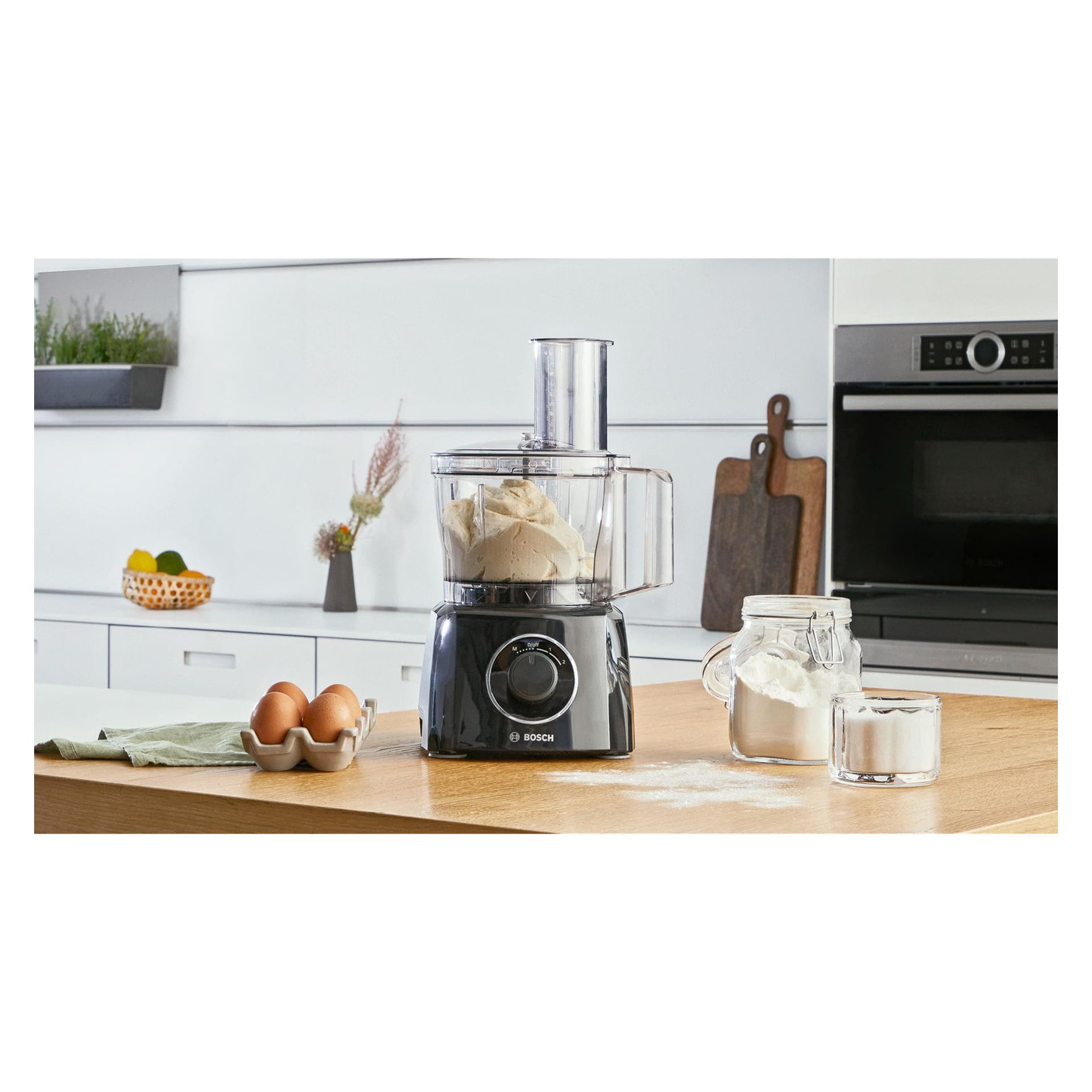 En Bosch MultiTalent 3 matprosessor med deig står på en hvit benk ved siden av tre egg, et glass med mel, sukker og et grønt tøystykke.