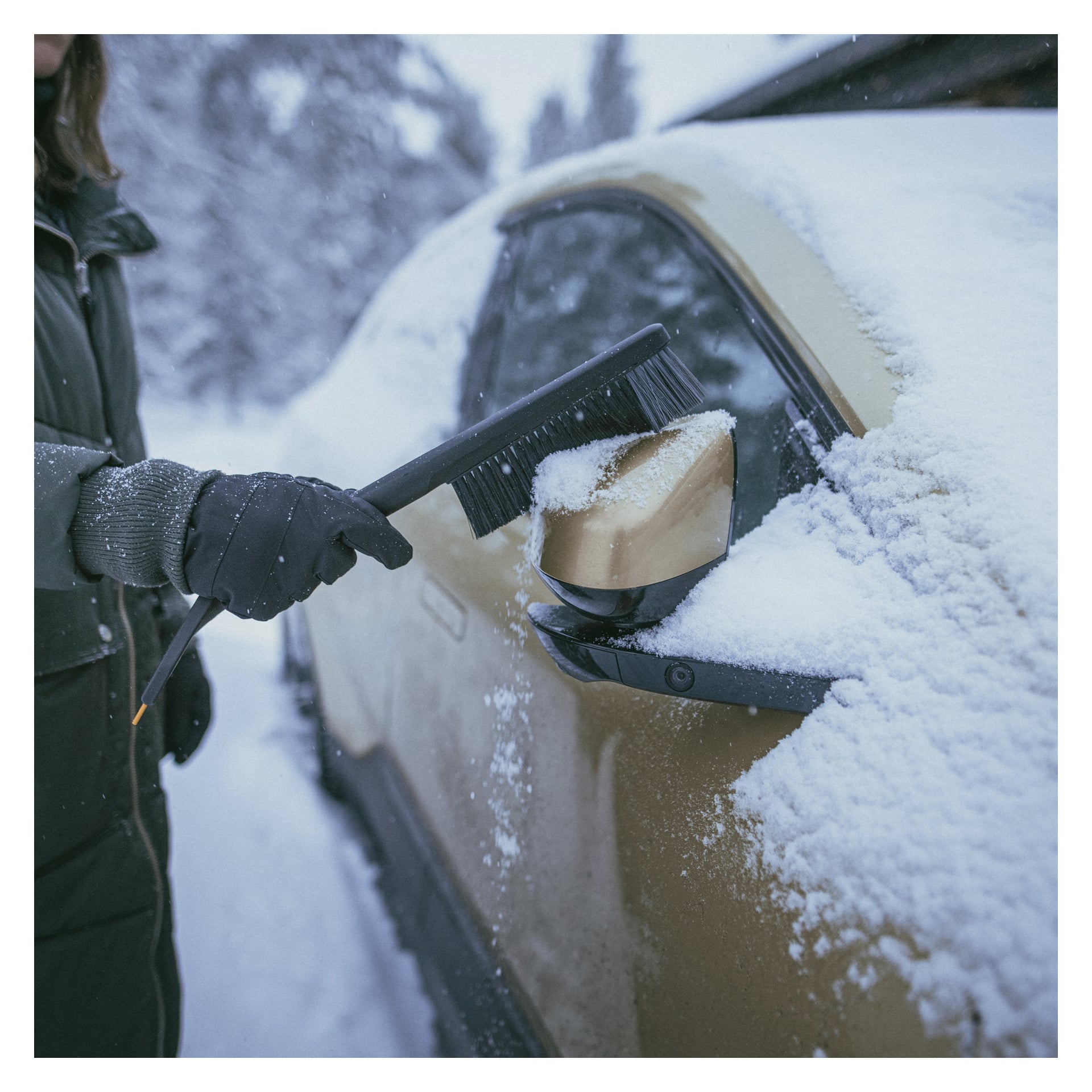Handskbeklädda händer använder Fiskars snöborste med isskrapa Solid för att ta bort snö från bilens sidospegel och fönster utomhus.
