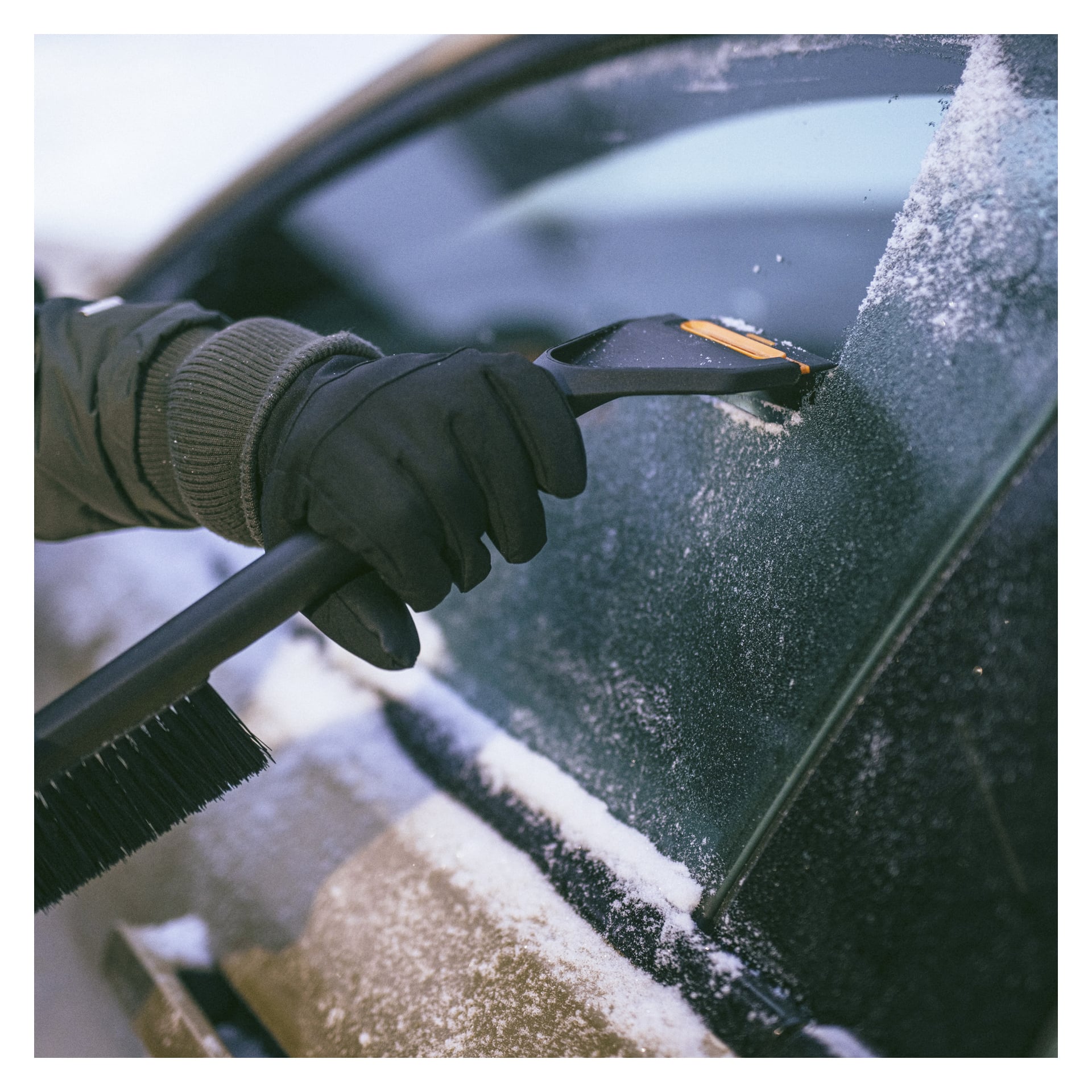 En person med handske använder en Fiskars Snow-borste med isskrapa för att ta bort frost från ett bilfönster täckt av snö och is.
