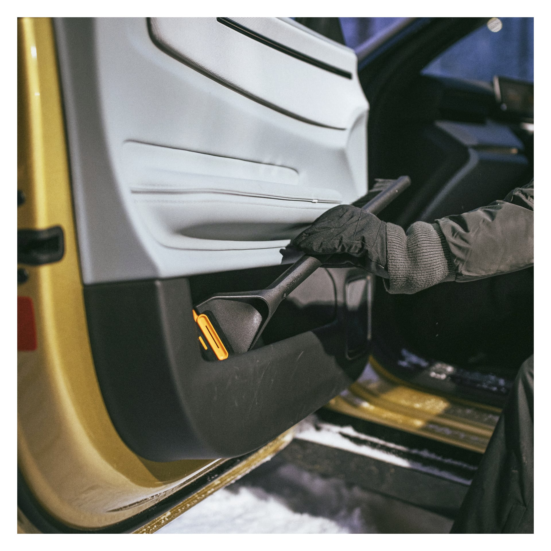 En handskbeklädd hand drar fram en Fiskars-snöbuske med orange handtag och isskrapa från en bildörrsglugg, med snö synlig utanför.