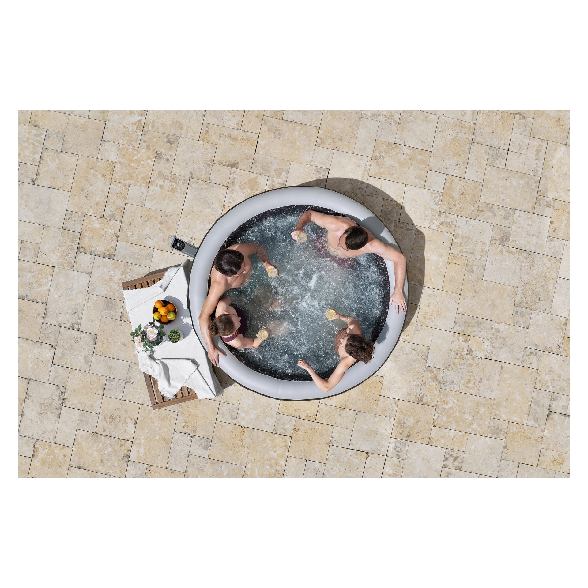 Fyra personer kopplar av i en rund bubbelpool på en uteplats av sten med drinkar i handen. Bredvid dem står ett bord med frukt, en flaska och en trasa.