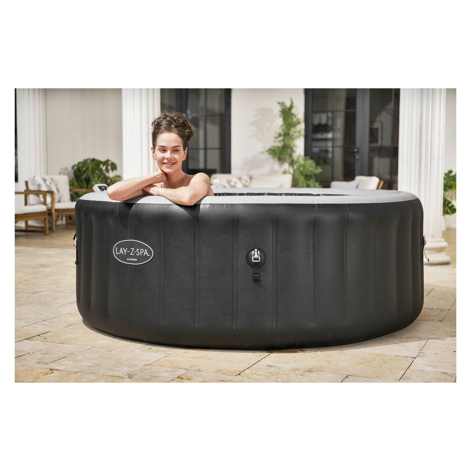 En person med mörkt hår i en uppsättning kopplar av i en svart Lay-Z-Spa-badtunna på en stenuteplats med vita pelare och utemöbler.
