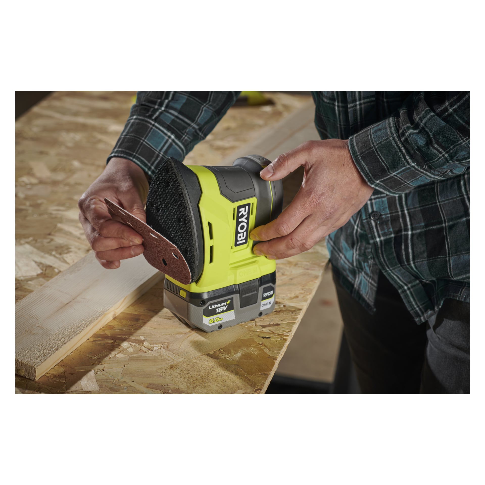 Man fäster ett Ryobi One+ SCS10A sandpappersset (10 stycken) på en sladdlös slipmaskin på en plywoodbänk nära en träplanka.