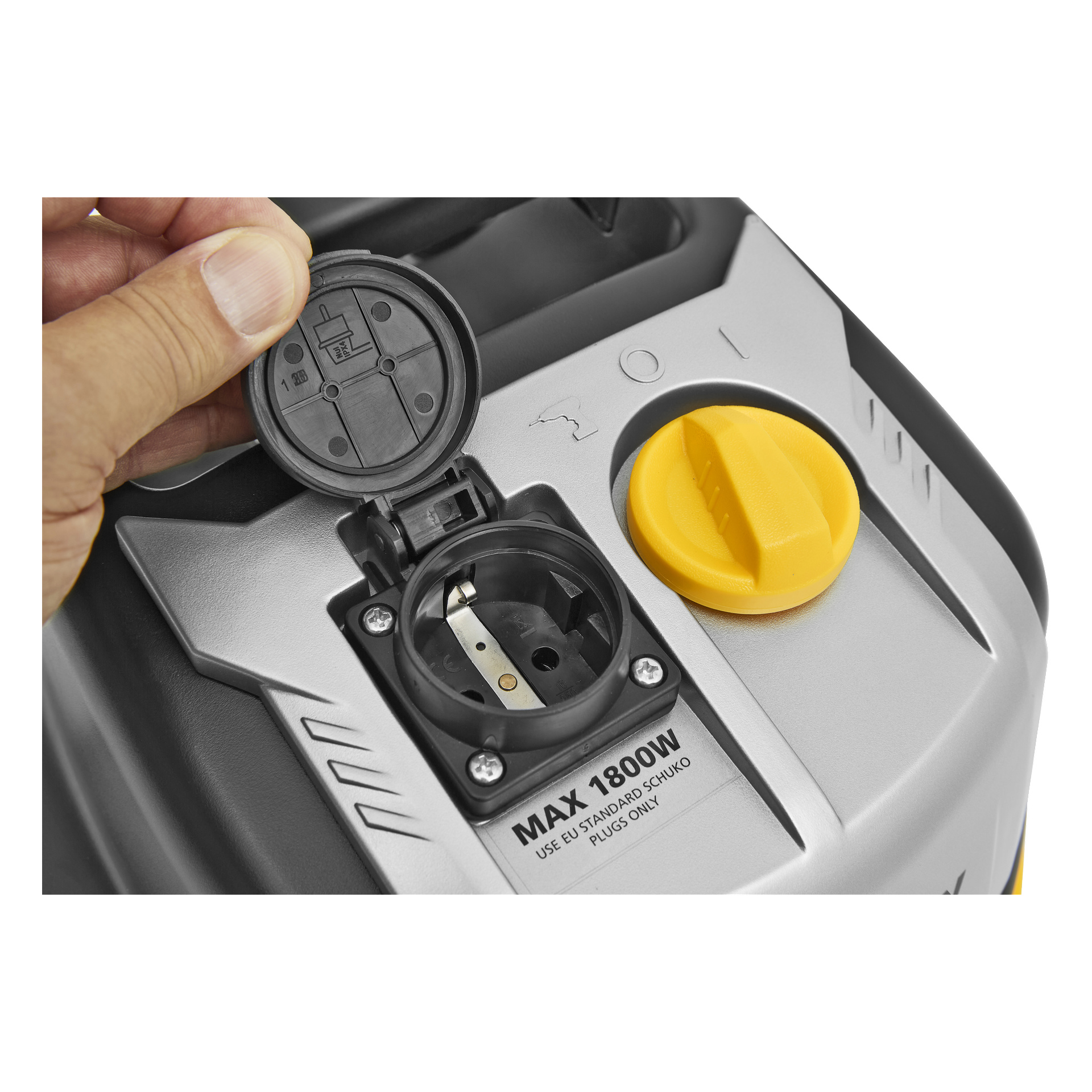 En hand lyfter på locket till ett svart eluttag på en silverfärgad och svart apparat märkt "MAX 1800W". En stor gul urtavla är placerad bredvid uttaget.