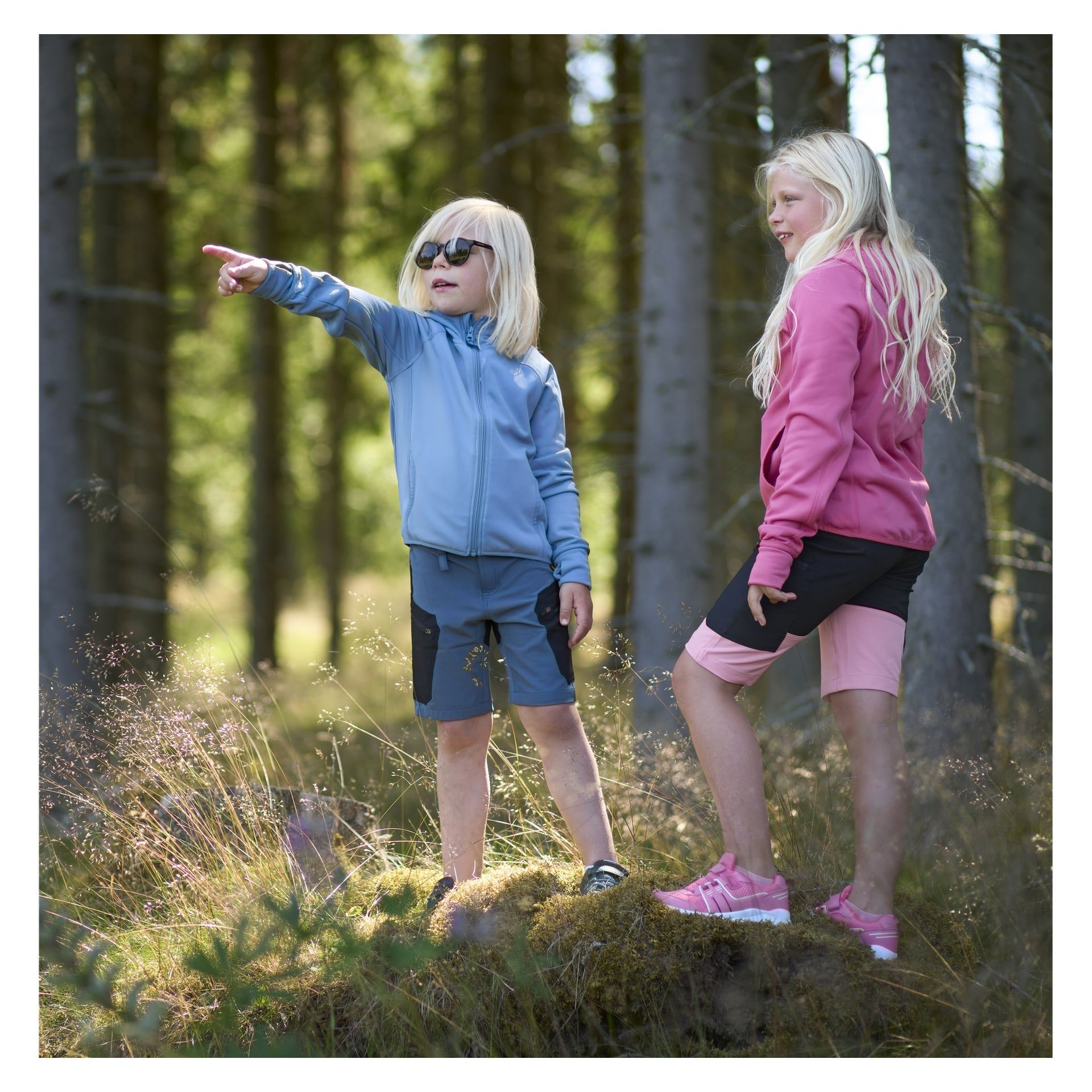 Två flickor, ena klädd i blåa och den andra klädd i rosa fritids shorts och tröja står på en lite kulle i skogen. Ena flickan pekar på något.