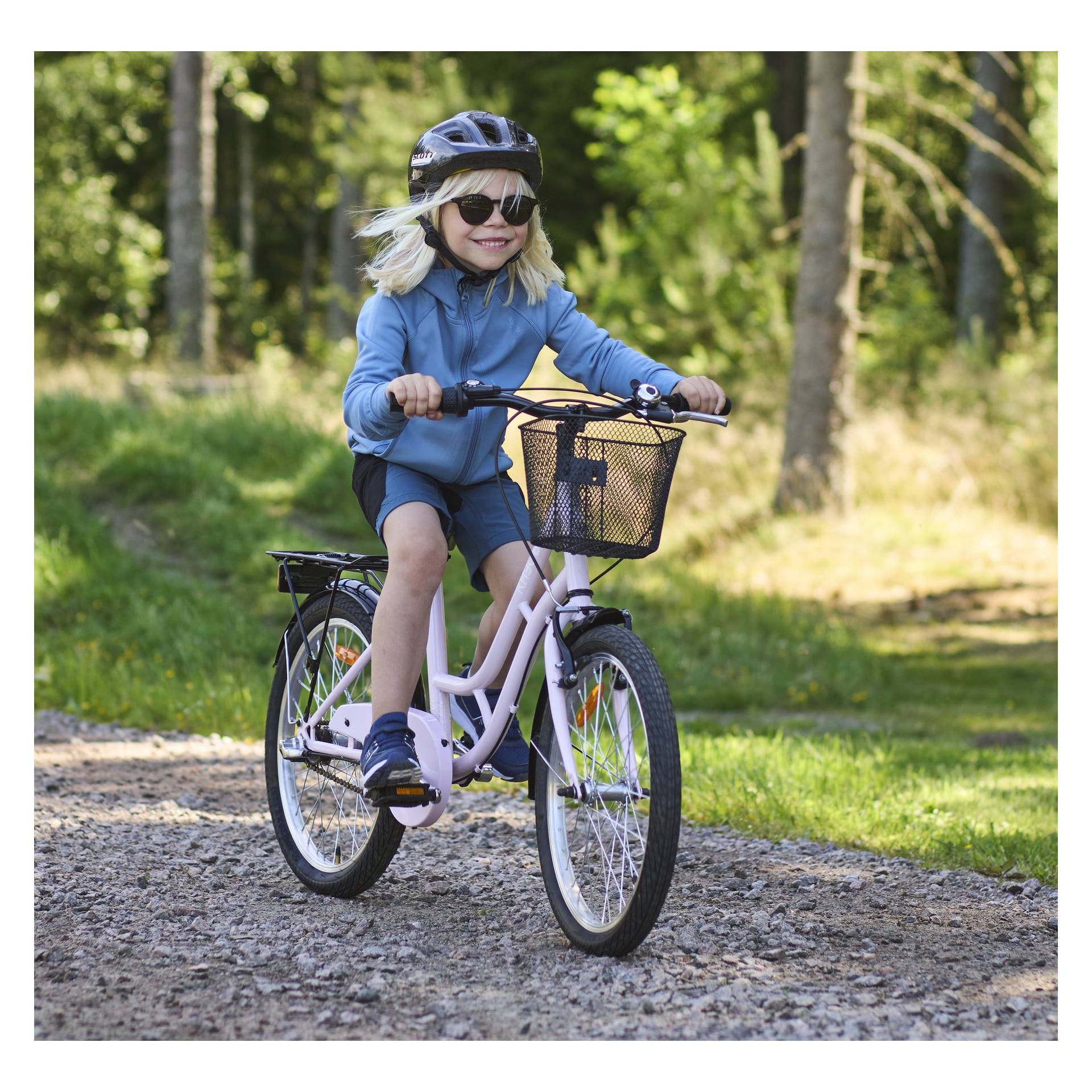 Flicka cyklar en rosa flickcykel på skogsväg. Hon har solglasögon, cykelhjälm och blå fritidskläder på sig.\n