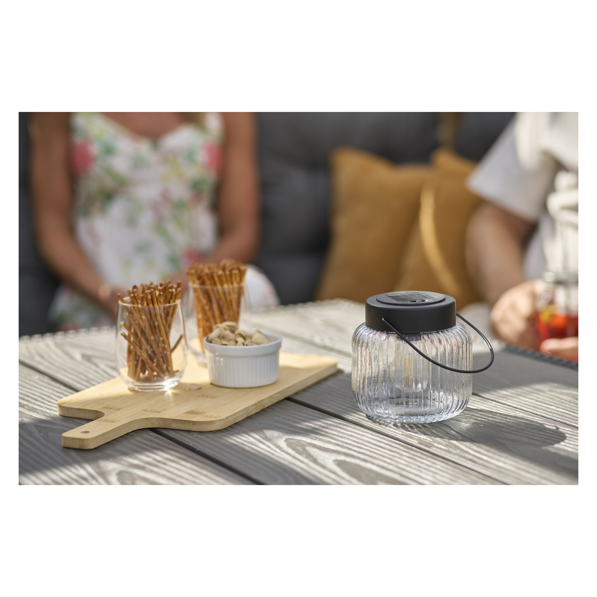 En Anslut LED Solar-lykt med svart lokk og håndtak står på et grått bord, snacks på en tavle og to uklare personer bak.