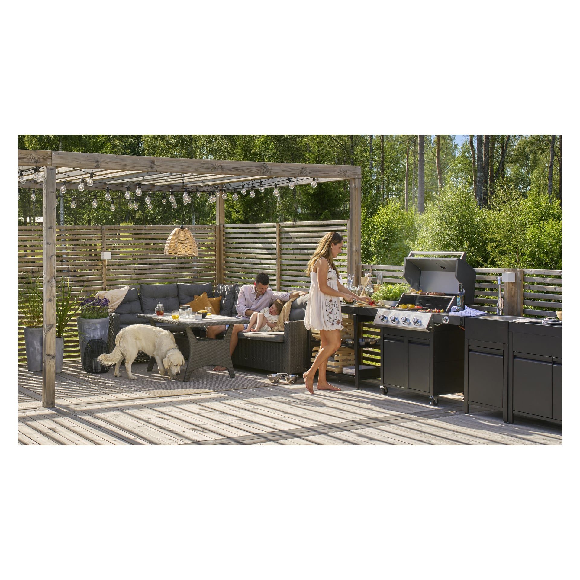 En kvinne griller på Outdoor Kitchen 8044B mens en mann og et barn slapper av i nærheten; En hvit hund snuser på terrassen blant grøntområdene.