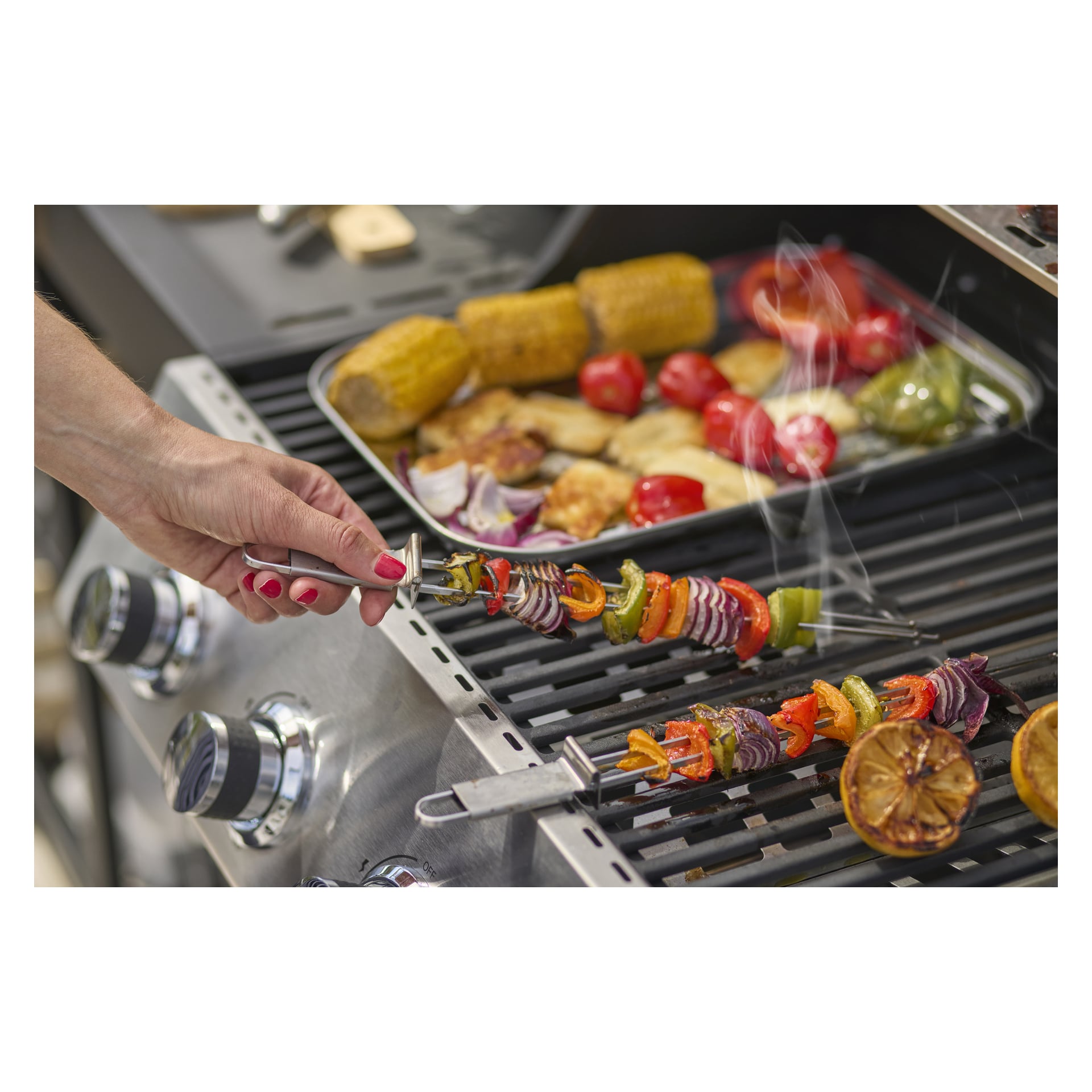 En hand håller ett Burns & Barkles Skewer 4-pack ovanför en grill fylld med spett, majs, tomater och grönsaker, med en bricka med mer mat.