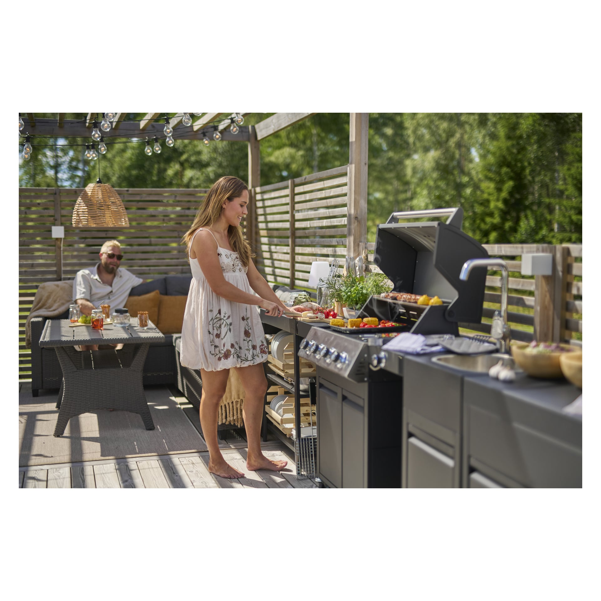 En kvinne griller på et utendørskjøkken på Burns & Barkles mens en mann slapper av i nærheten på en solrik terrasse med lysslynger og grøntområder.