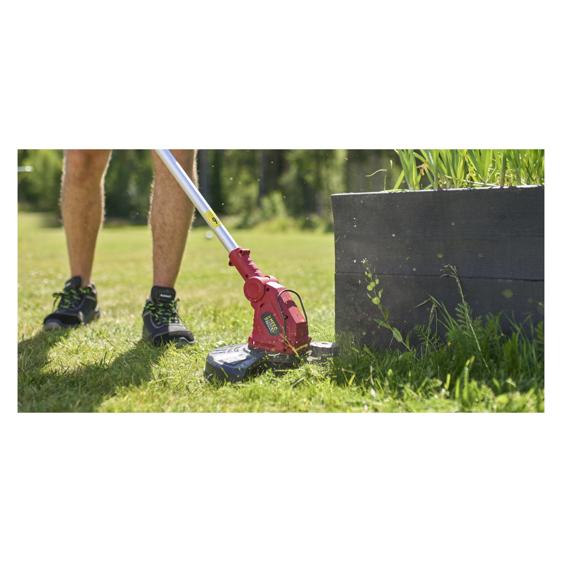 En person i svarta sneakers klipper gräs bredvid en träkruka med en Meec Tools Multiseries GT-18V HOME 500 batteritrimmer.