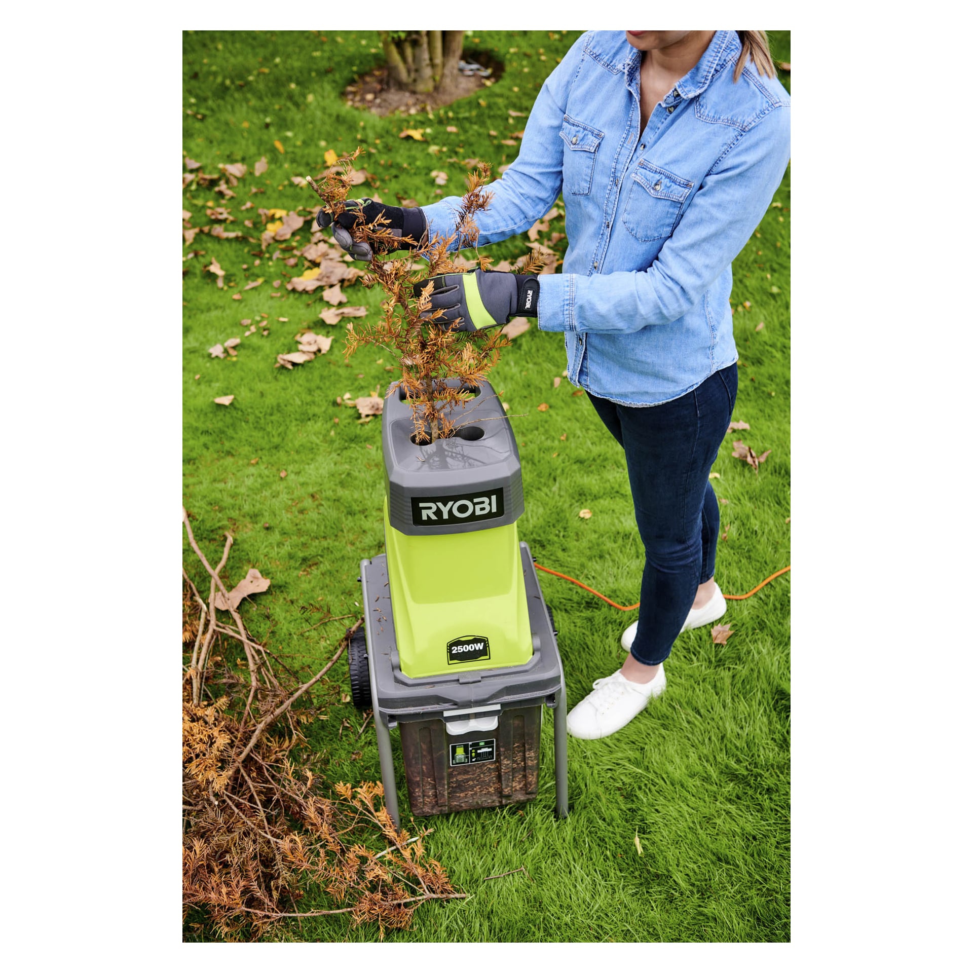 En person i blå skjorte og hansker mater tørre greiner inn i en grønn og grå Ryobi-makulator på en plen med spredte blader.