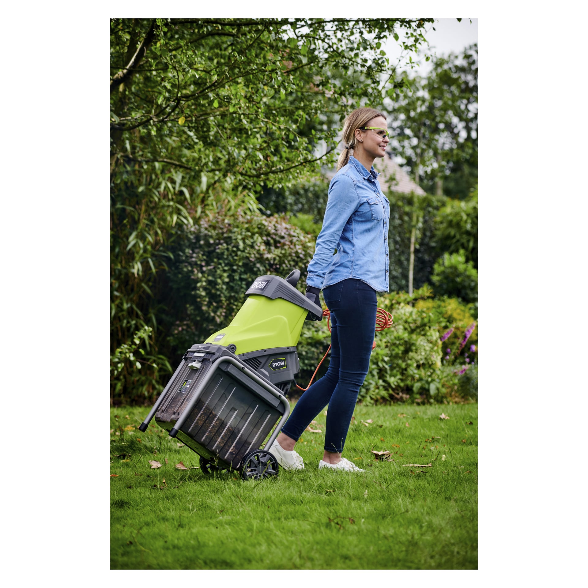 Iført en blå dongeriskjorte og mørke jeans drar en person en Ryobi Compost Shredder RSH2545B over gresset i en utendørs hage.