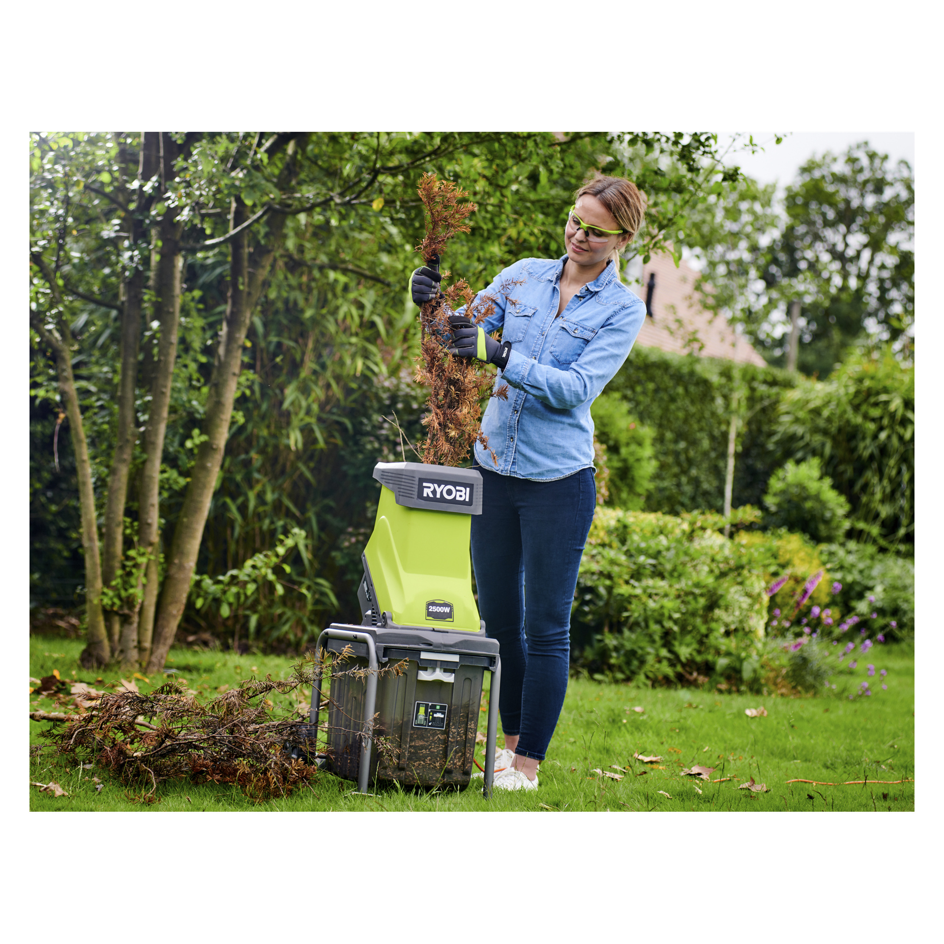 En person med vernebriller og hansker mater tørkede greiner inn i en Ryobi Compost Shredder RSH2545B i en gresshage med trær.