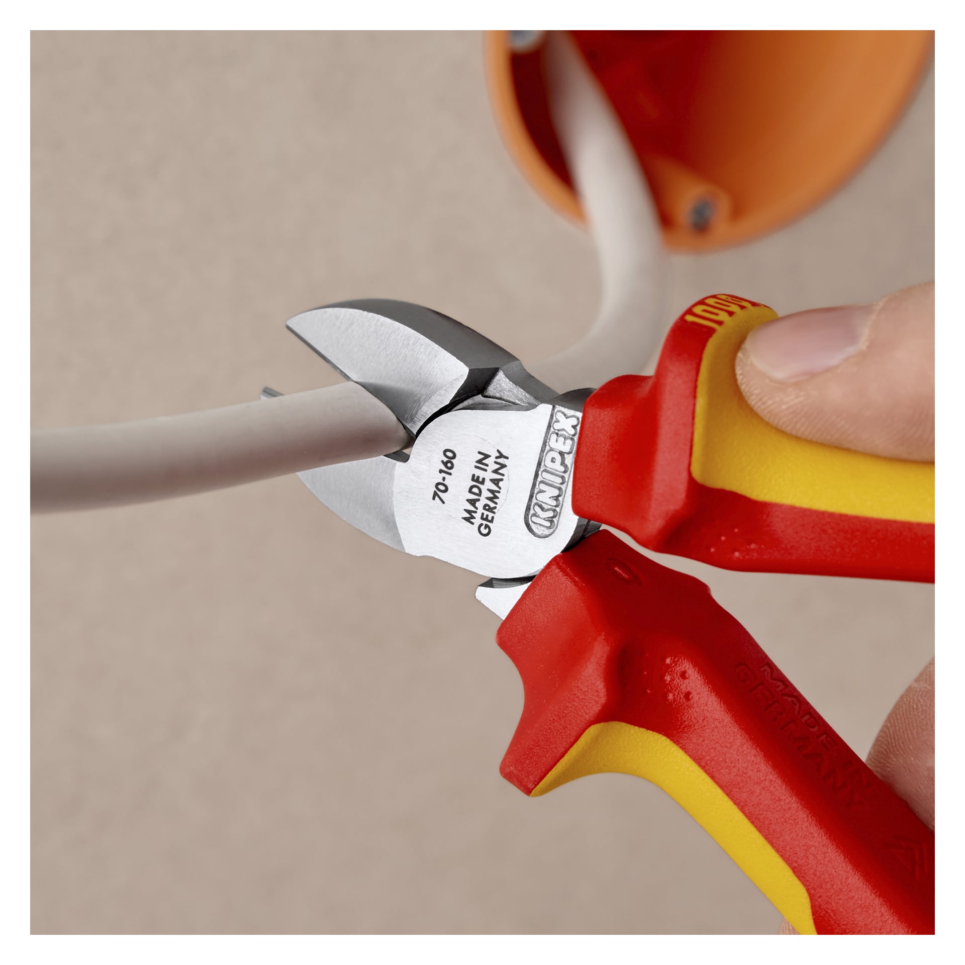 Ręka obsługuje czerwone i żółte szczypce Knipex do cięcia bocznego, aby przeciąć gruby biały kabel elektryczny obok pomarańczowej puszki ściennej.