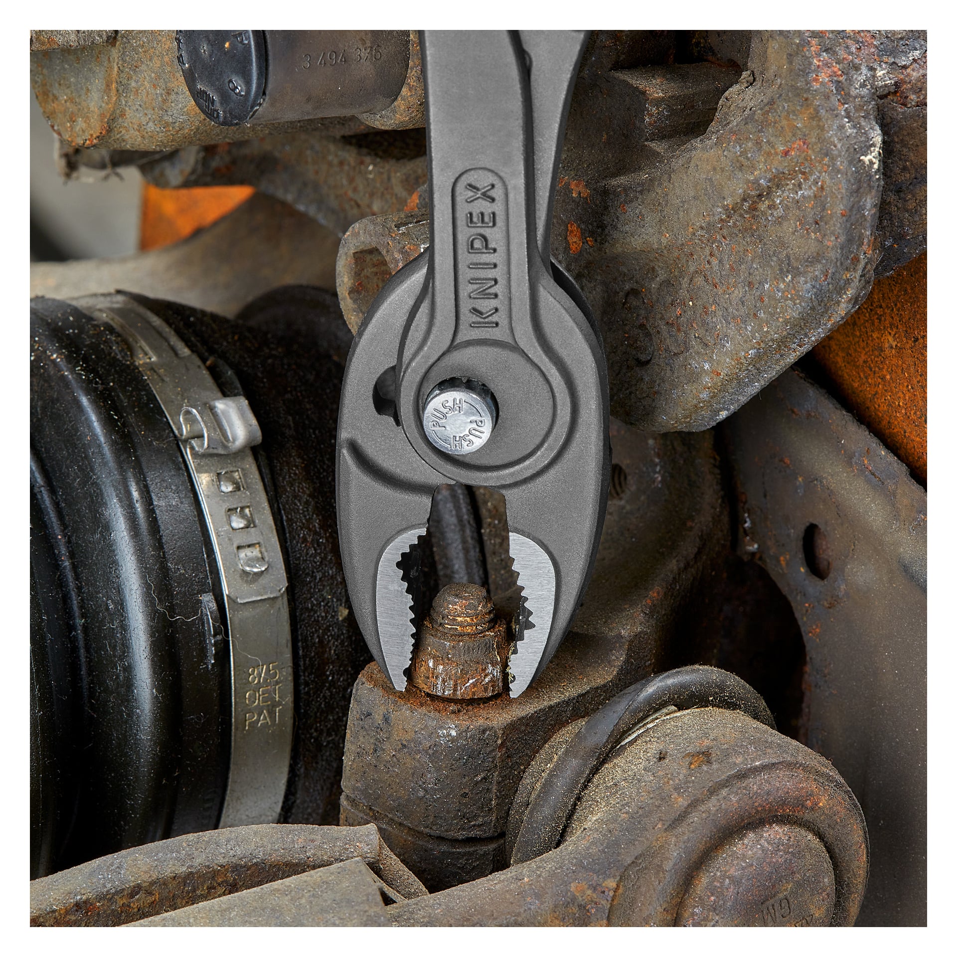 Knipex TwinGrip kombinasjonstenger holder sikkert en rusten, avrundet bolt blant slitte metalldeler i bilmiljø.