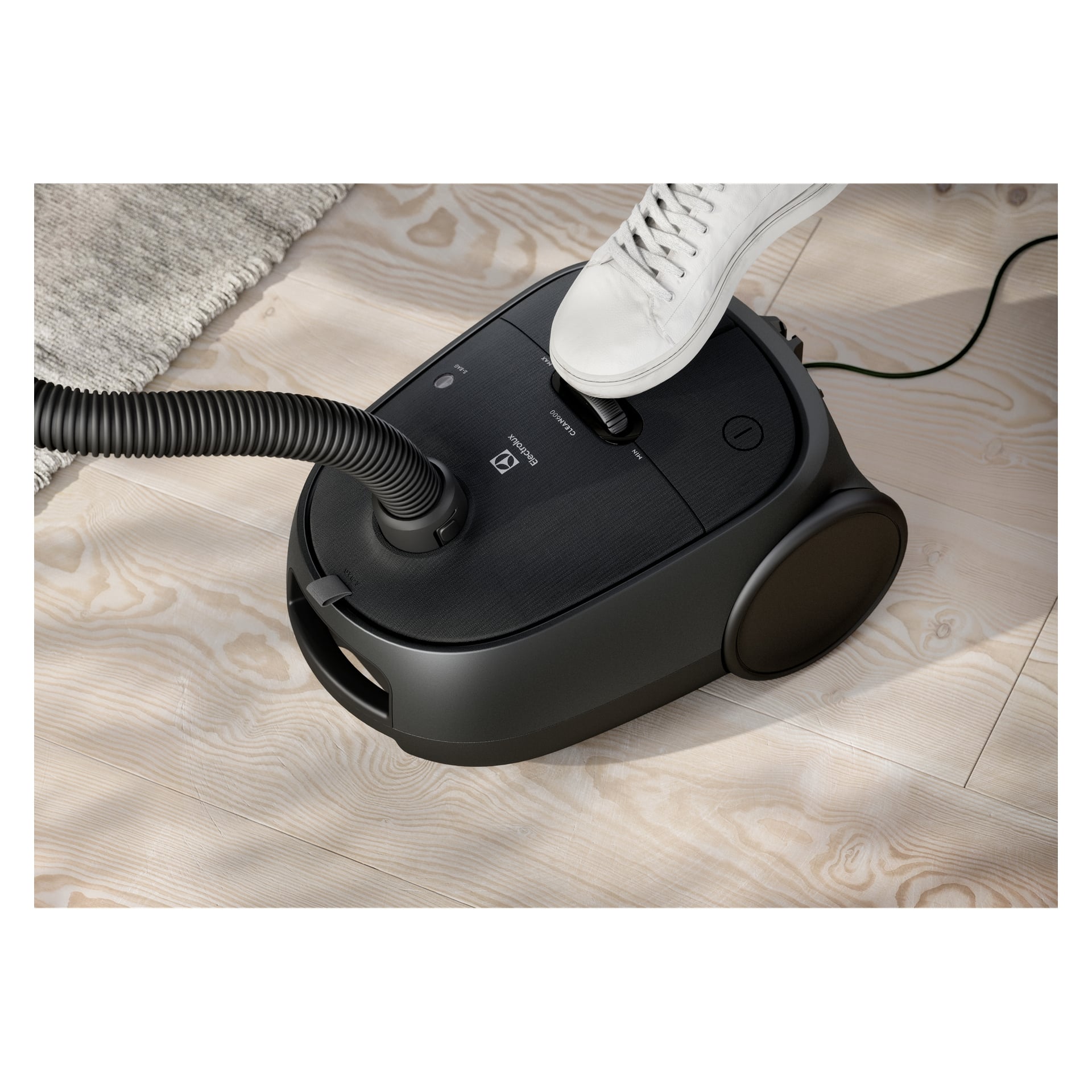Iklädd vita sneakers trycker man på strömknappen på en svart Electrolux Clean 600-dammsugare på ett lätt trägolv vid en matta.