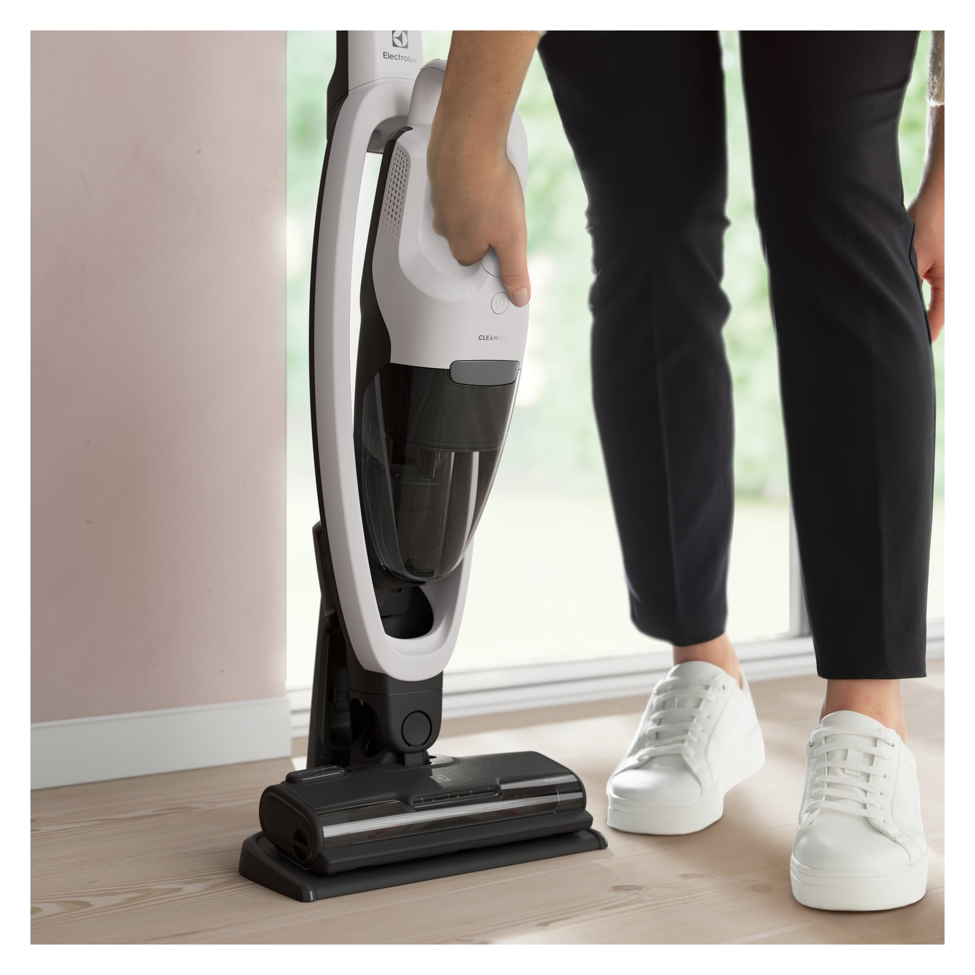 Iført svarte bukser og hvite joggesko løsner en person håndholdt enhet fra en Electrolux Upright støvsuger på et lyst tregulv ved et vindu.