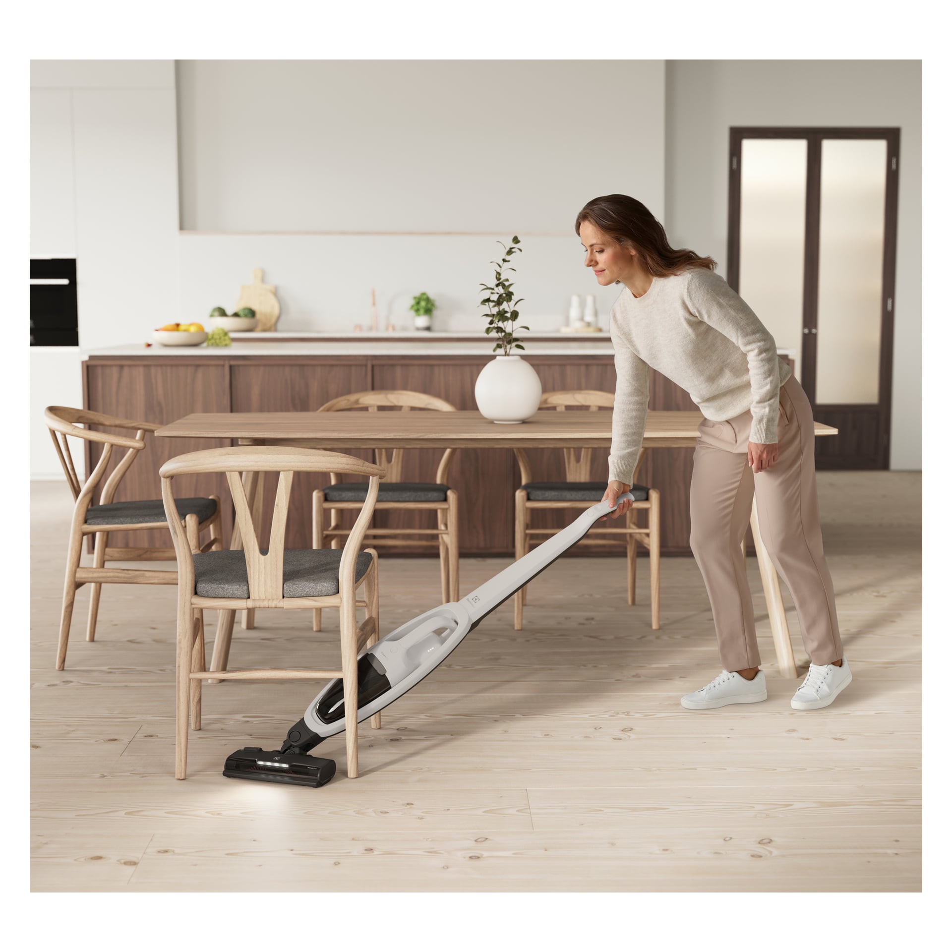 En uformelt kledd kvinne støvsuger et lyst tregulv i en moderne spisestue med trebord og stoler ved hjelp av en Electrolux 500 Clean.