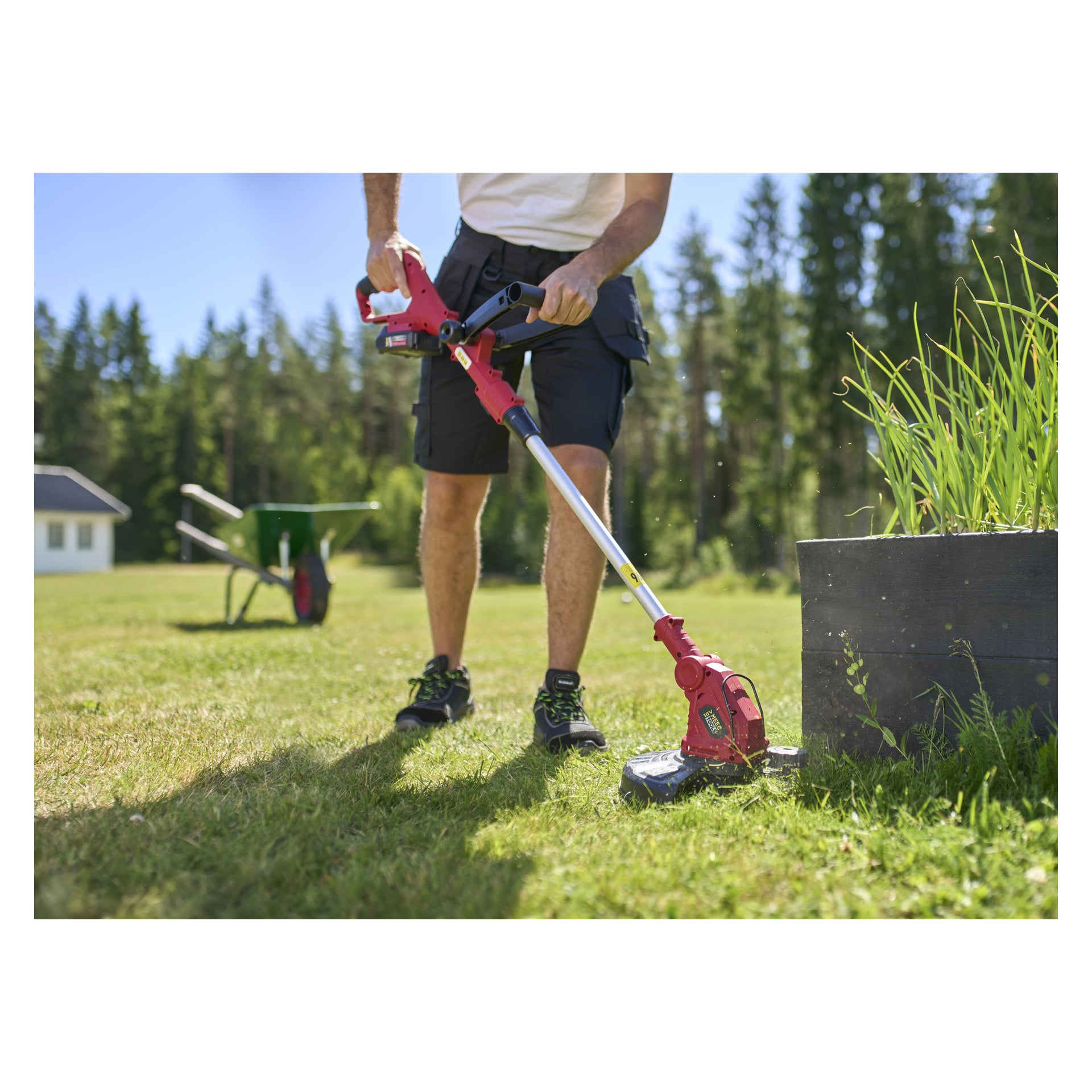 Henkilö leikkaa nurmikkoa korotetun puutarhapenkin vieressä käyttäen Meec Tools Multiseries -sarjasarjaa; Puut ja vihreä kottikärry ovat auringonvalossa taustalla.