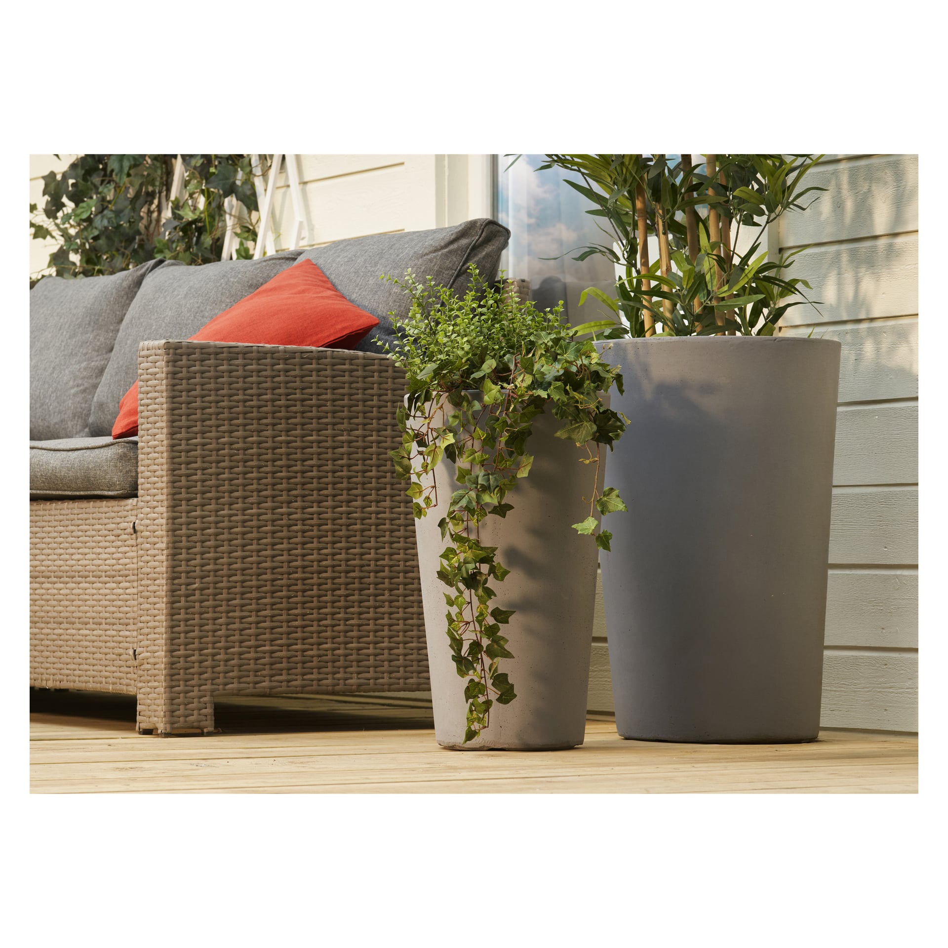 To høye potteplanter i Hard Head-potter på en treterrasse ved siden av en kurvsofa med grå pute og rød pute ved en lysvegg.