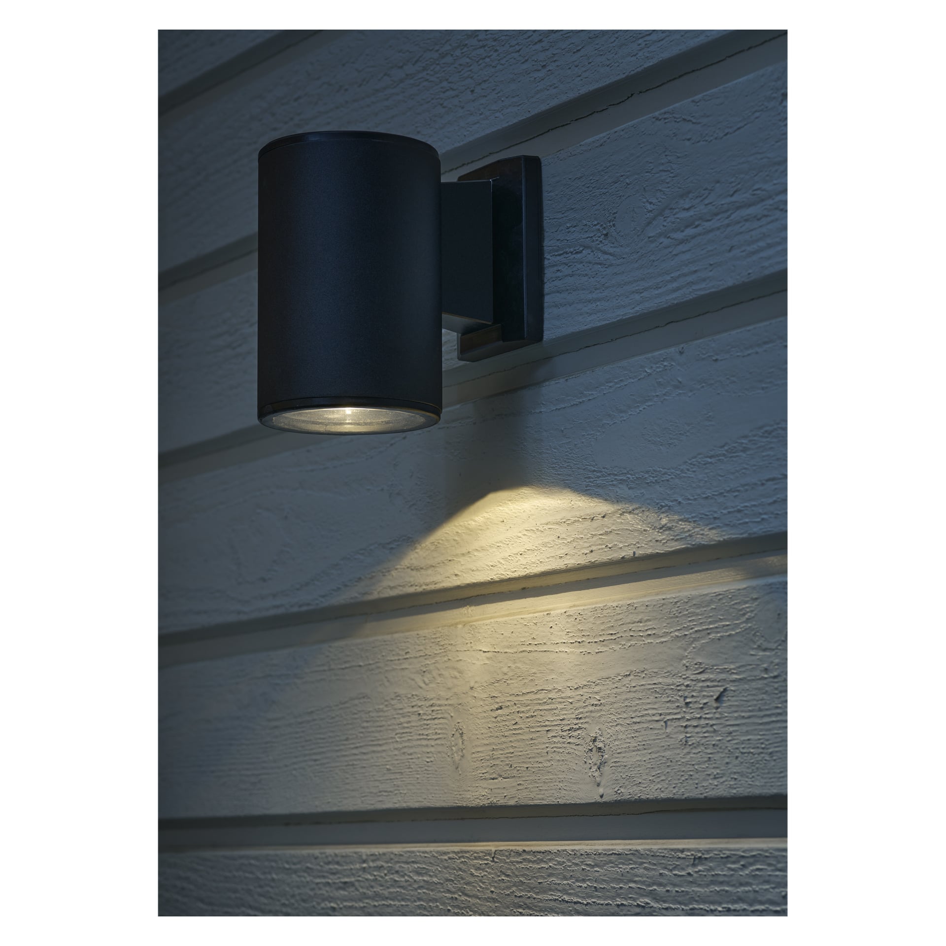 Svart nedåtriktad solcellsdriven vägglampa lyser upp den vita väggen i mörkret.
