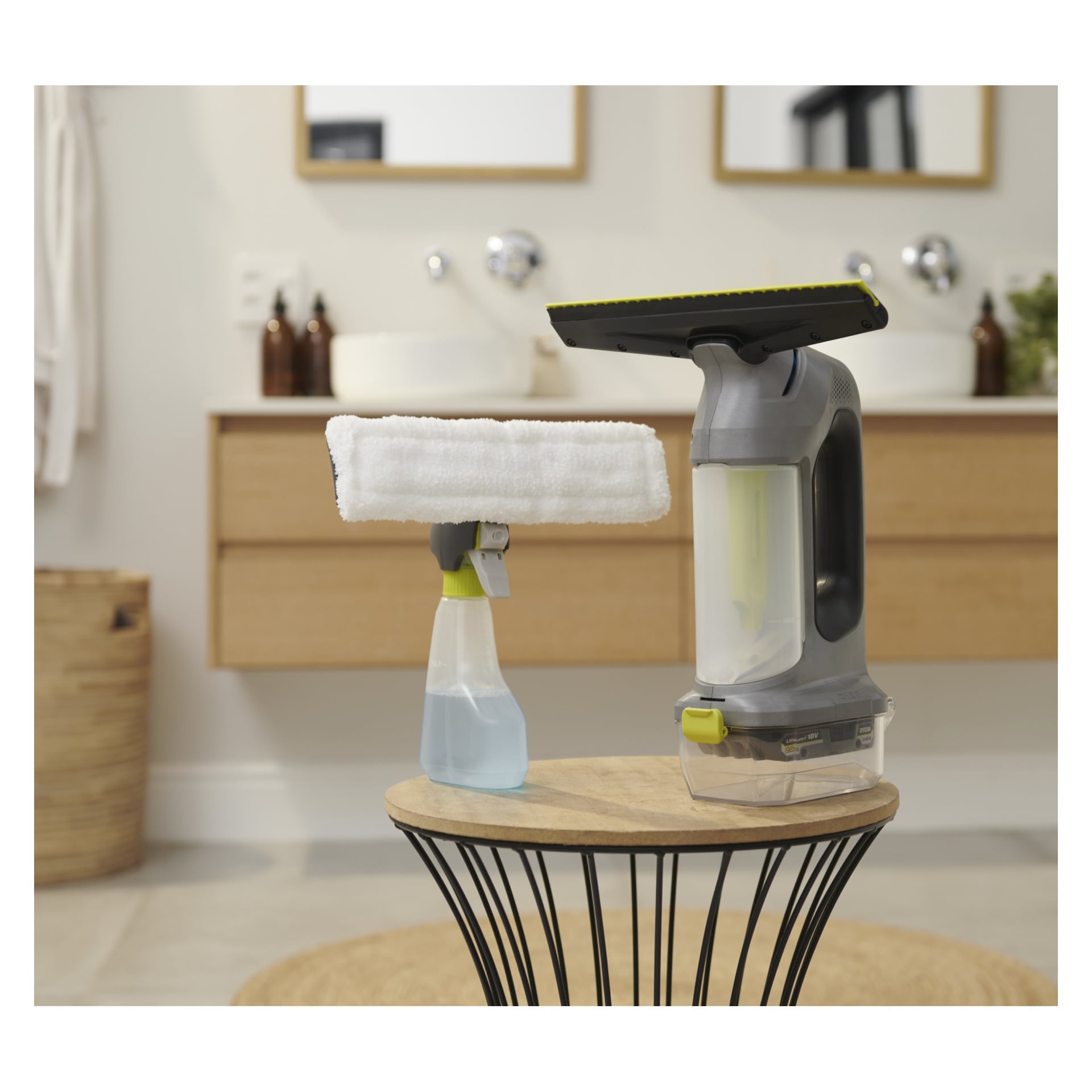 En Ryobi One+ sladdlös fönsterputsare och en sprayflaska med mikrofiberdyna vilar på en träpall i ett modernt badrum med ljusa träskåp och speglar.