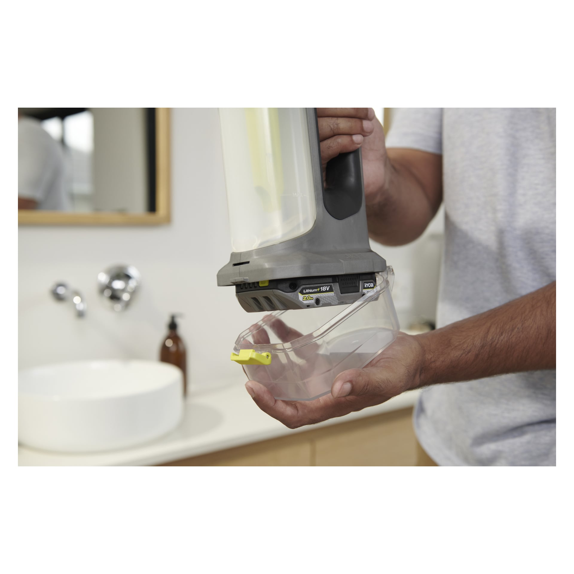 Någon lossar den genomskinliga plastbehållaren från en Ryobi One+ trådlös fönsterputsare; En handfat, kran och tvålautomat syns.