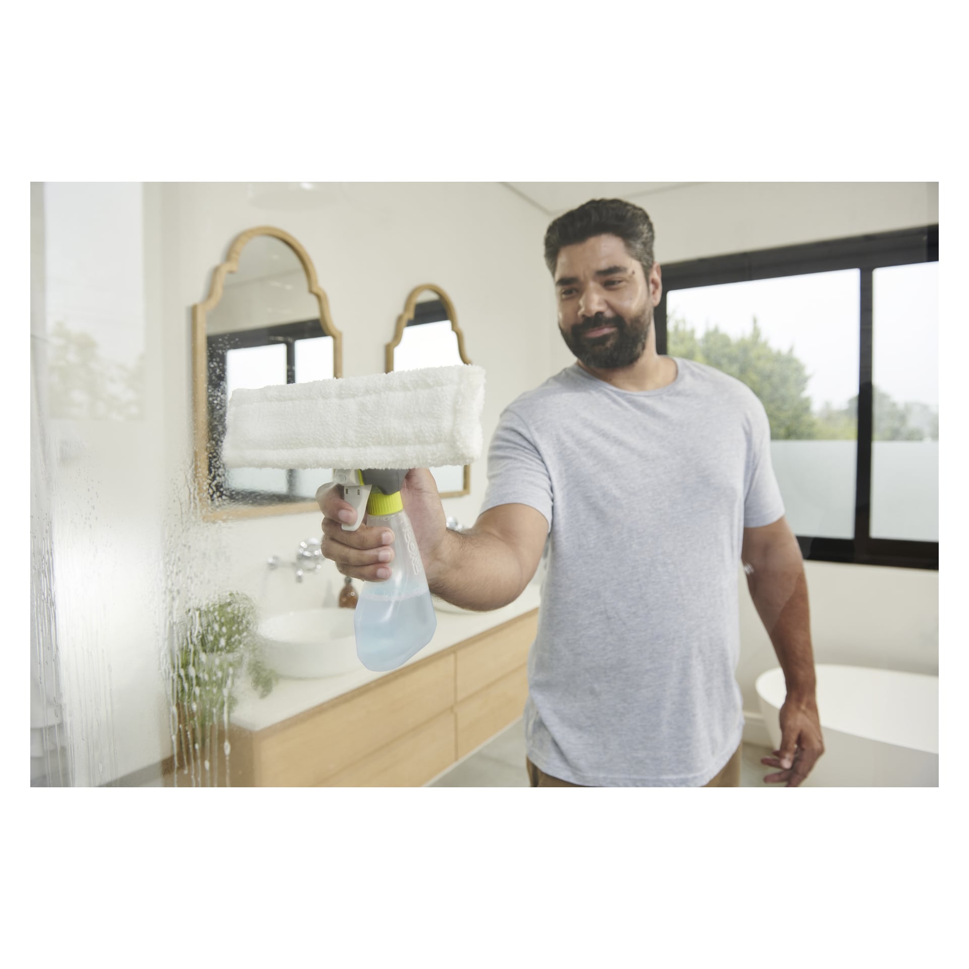 En man i grå T-shirt rengör en glasdörr till duschen med Ryobi One+ Cordless fönsterputsare i ett solbelyst badrum med träkommodiär och speglar.
