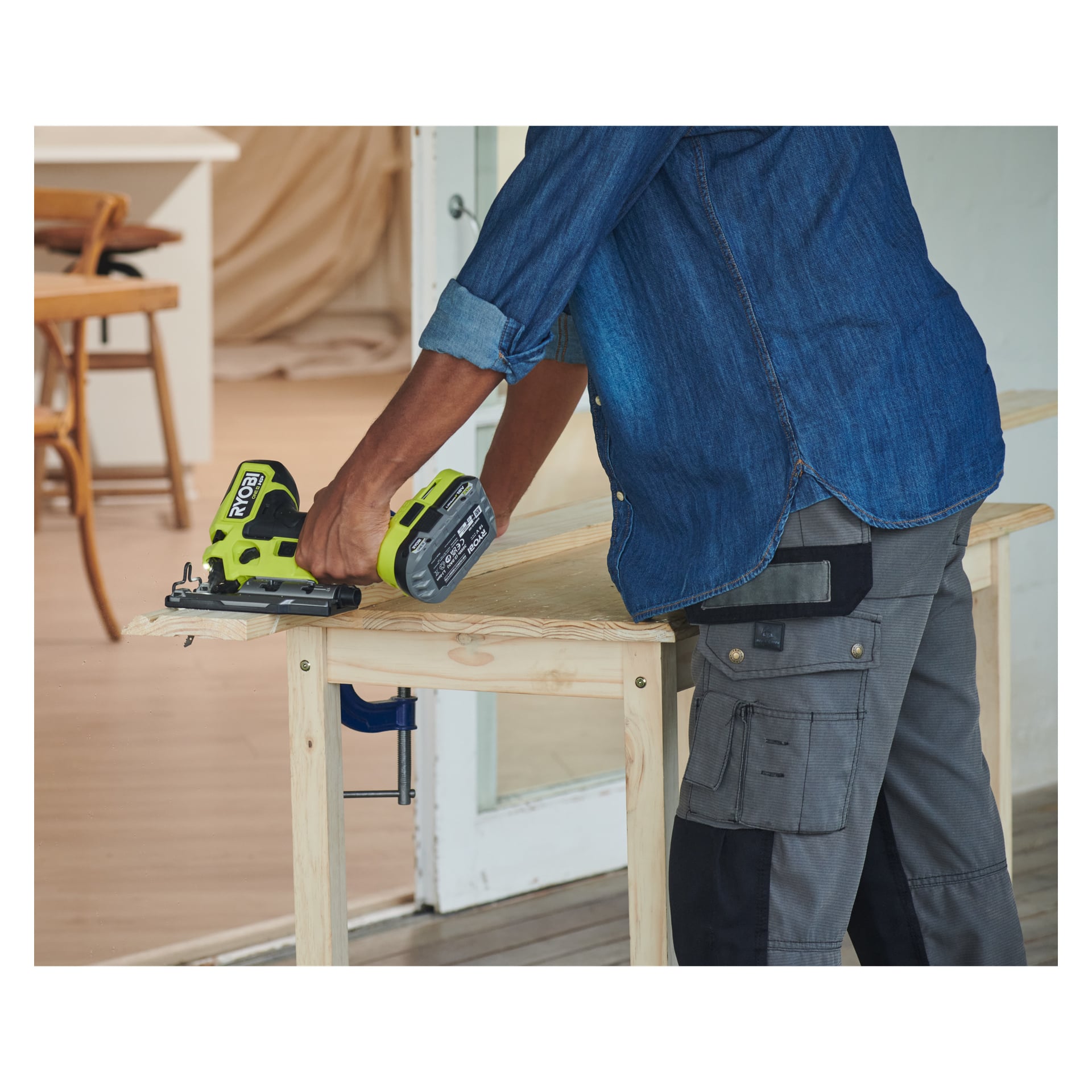 En person i jeansskjorta och arbetsbyxor använder en Ryobi One+ HP sticksåg för att såga en träskiva på en fastklämd arbetsbänk inomhus.